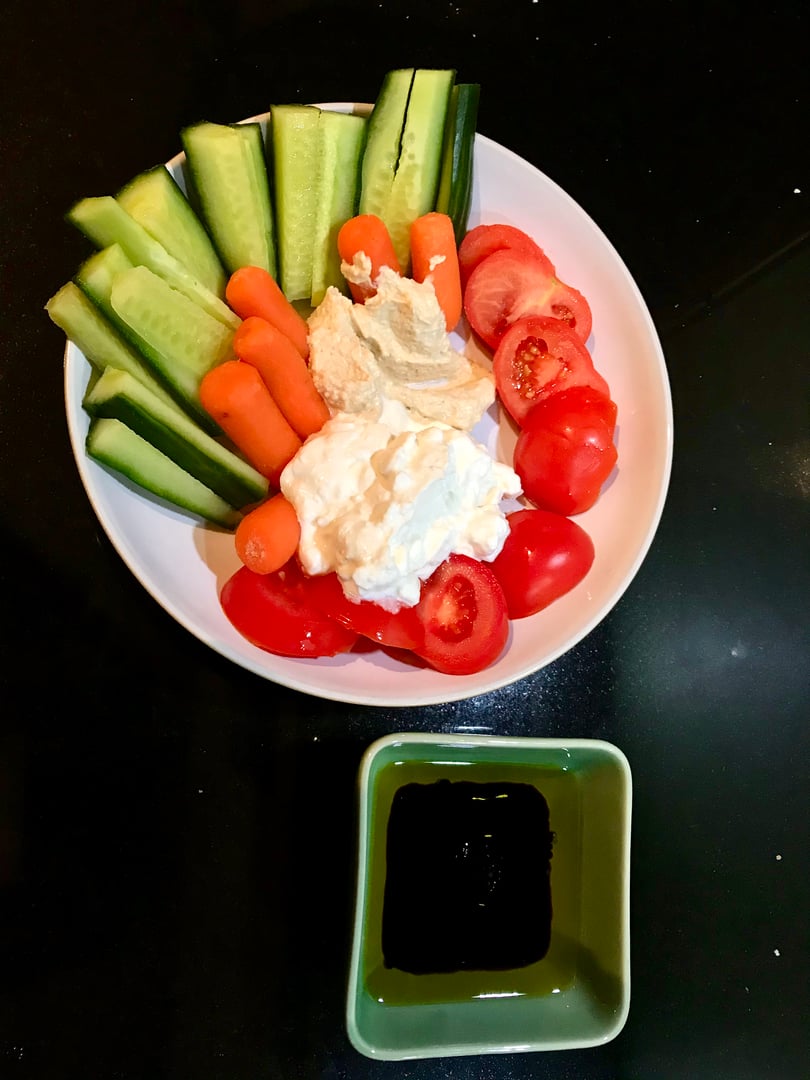 Homemade cucumber sticks, carrot sticks, baby tomatoes, dollop of cottage cheese and humous in white bowl with side dish of extra virgin olive oil and balsamic vinaigrette on black marble background