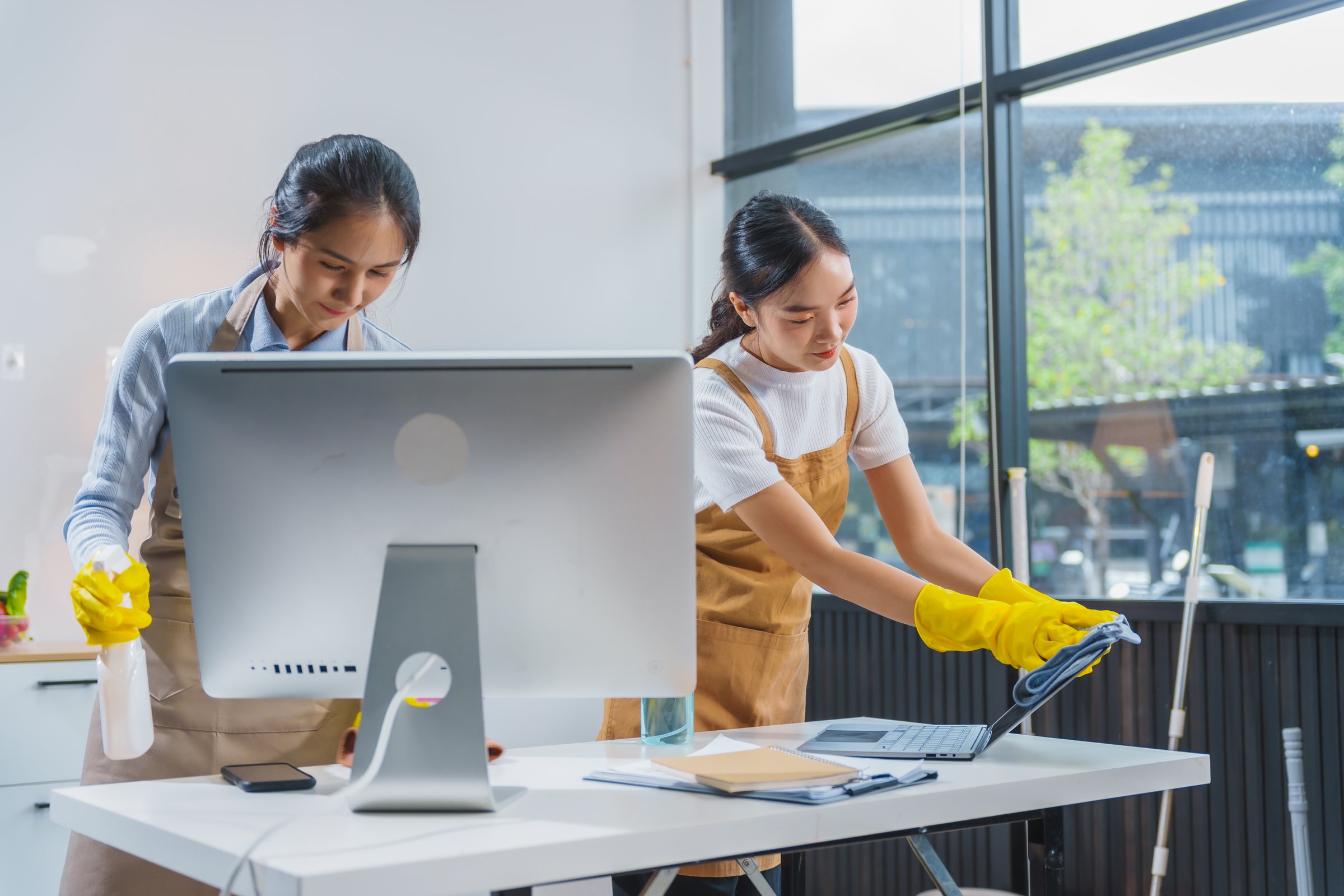 Two Asian housekeepers, wearing aprons and rubber gloves, happily clean an office. They sweep the floor, wipe glass surfaces, tidy up the sofa using cleaning liquids, a broom, and microfiber cloths.