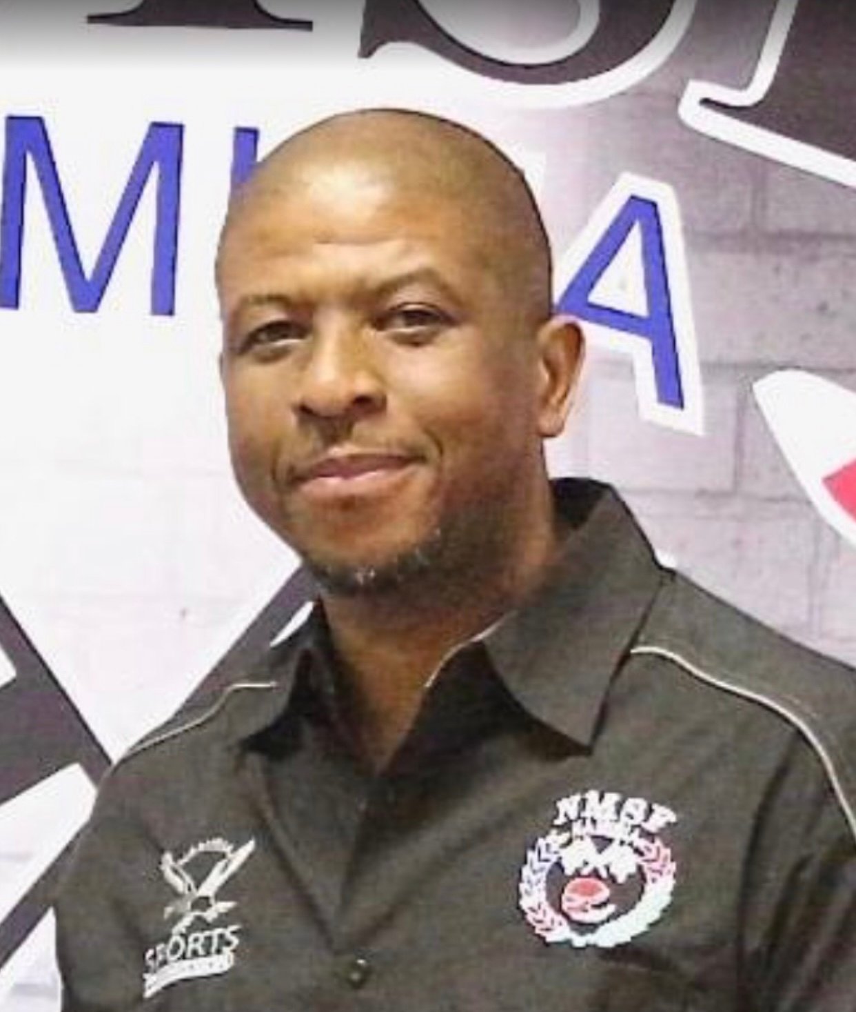 A man smiling, wearing a dark shirt with logos, stands in front of a partially visible mural or sign.