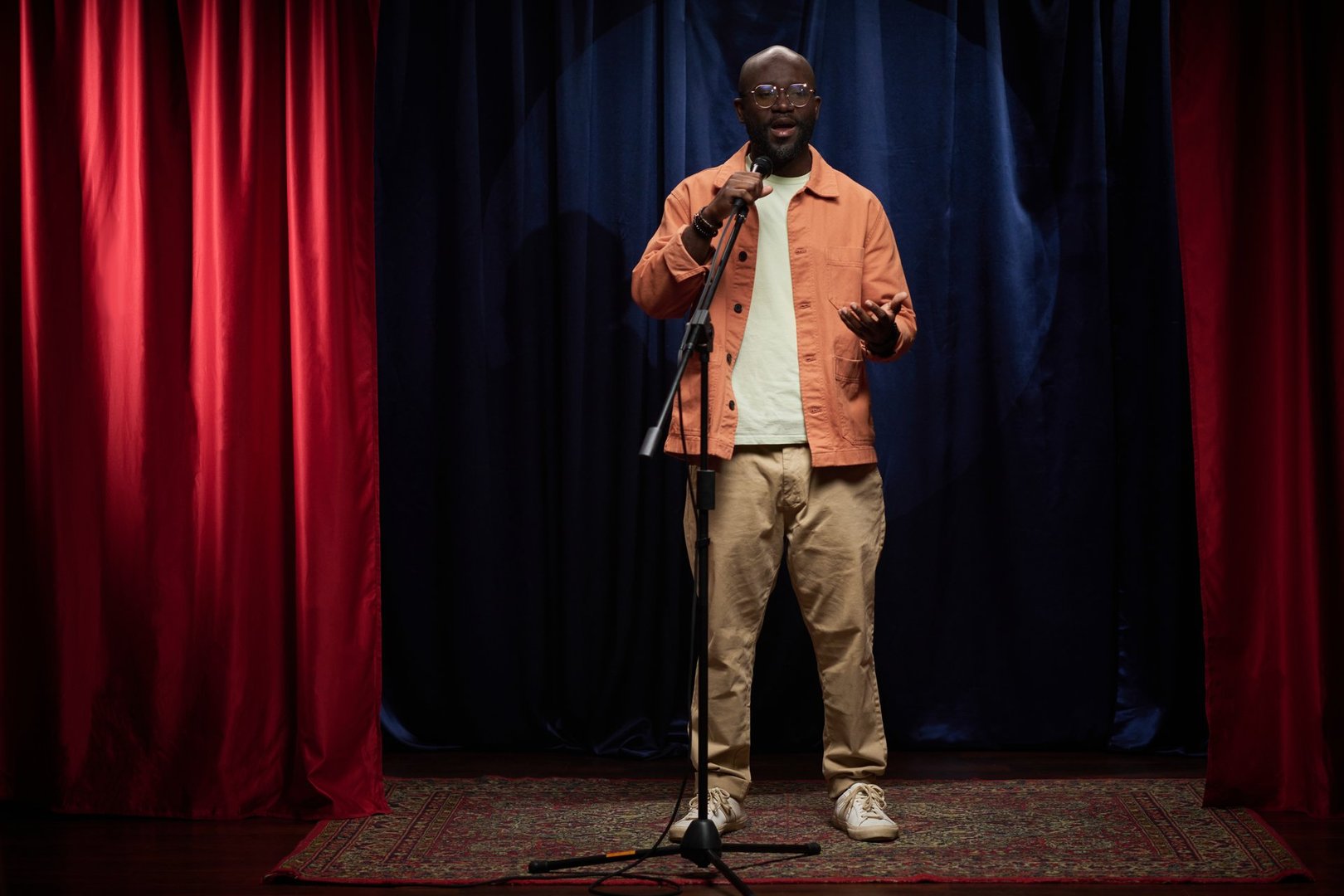 Person performing stand-up comedy on stage, engaging with audience while holding microphone, wearing casual outfit, standing on small stage with red curtains