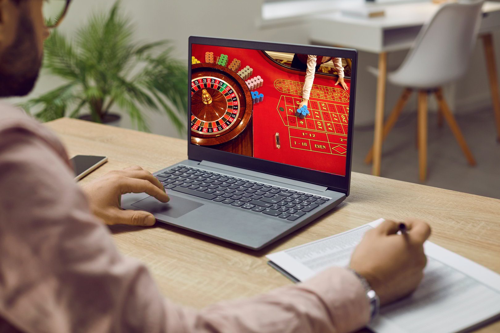 Man placing bets and writing notes while playing online casino at home on his laptop. Close up of laptop screen on which roulette is being played and which man addicted to gambling is looking at.