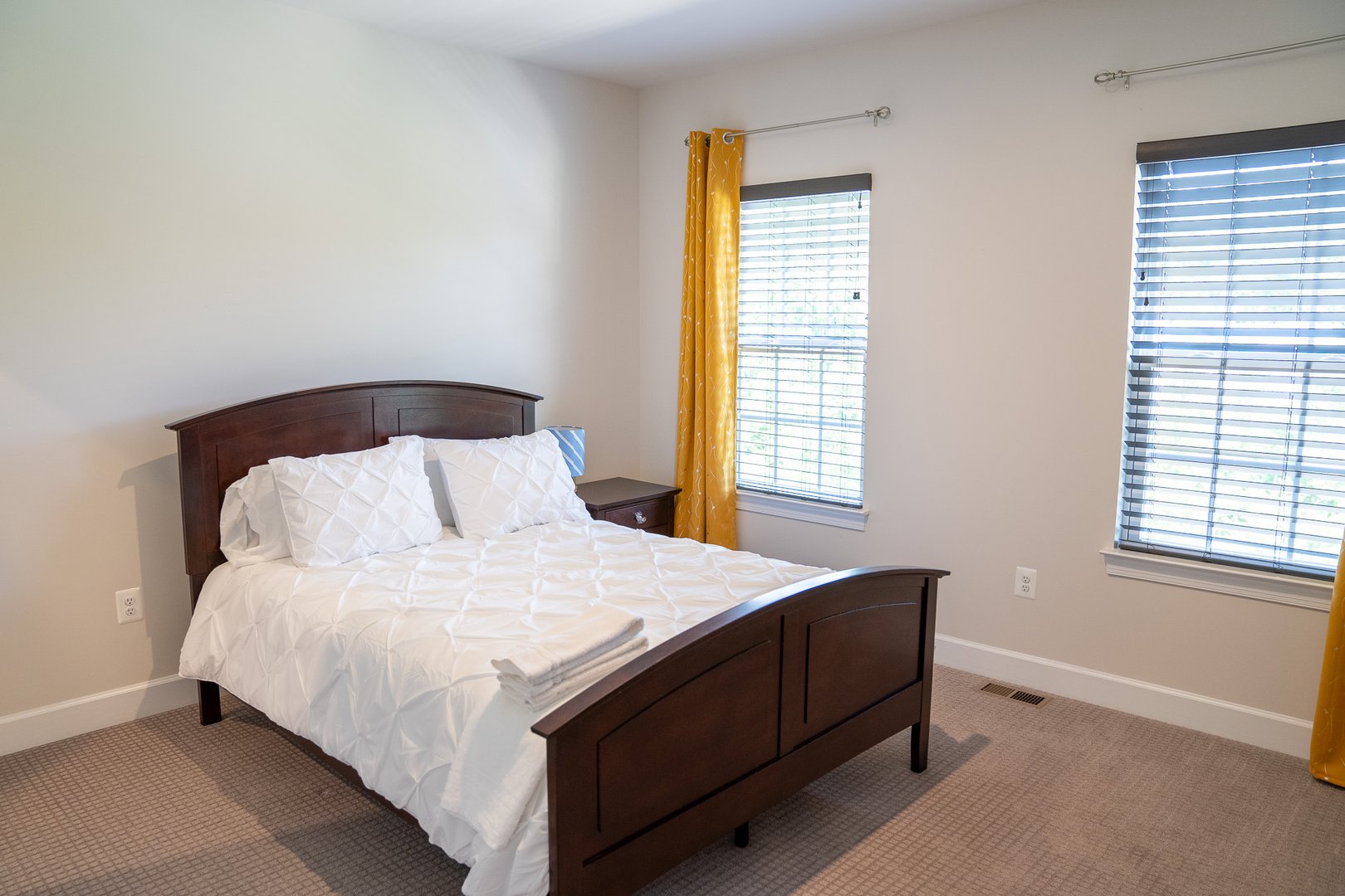 Bedroom with a wooden bed frame, white bedding, yellow curtains, and two windows with blinds letting in natural light.