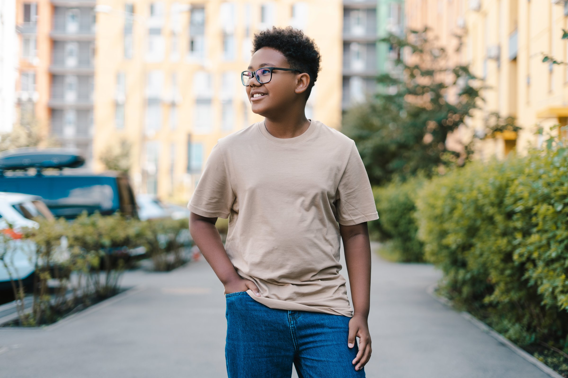 Confident young man standing on city sidewalk in summer day, wearing trendy beige t-shirt with copy space, stylish glasses, exuding happiness and youthfulness