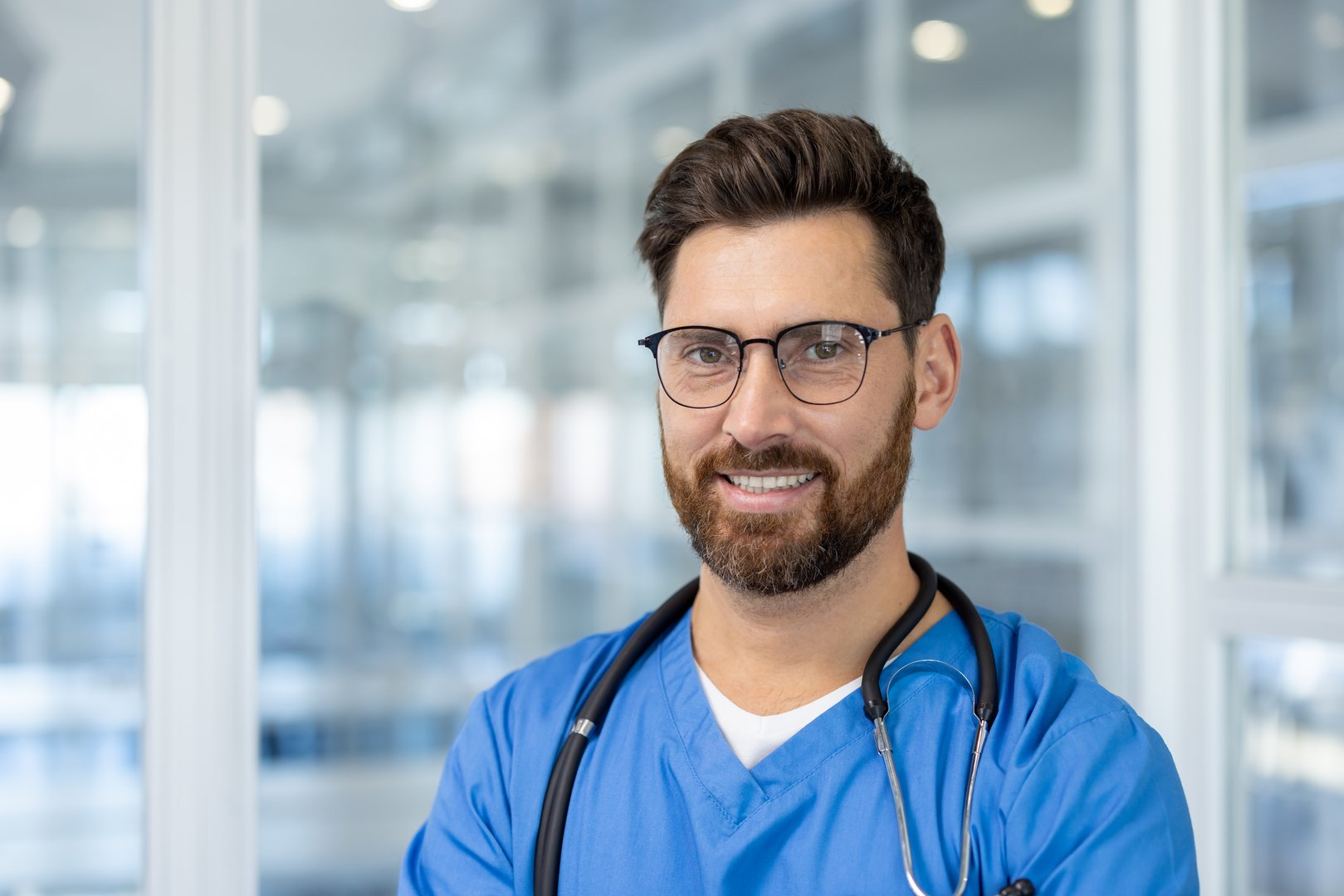 Young man doctor in blue scrubs and glasses in hospital corridor, exuding confidence and professionalism. Ideal for showcasing healthcare professional themes. Smiling with stethoscope.