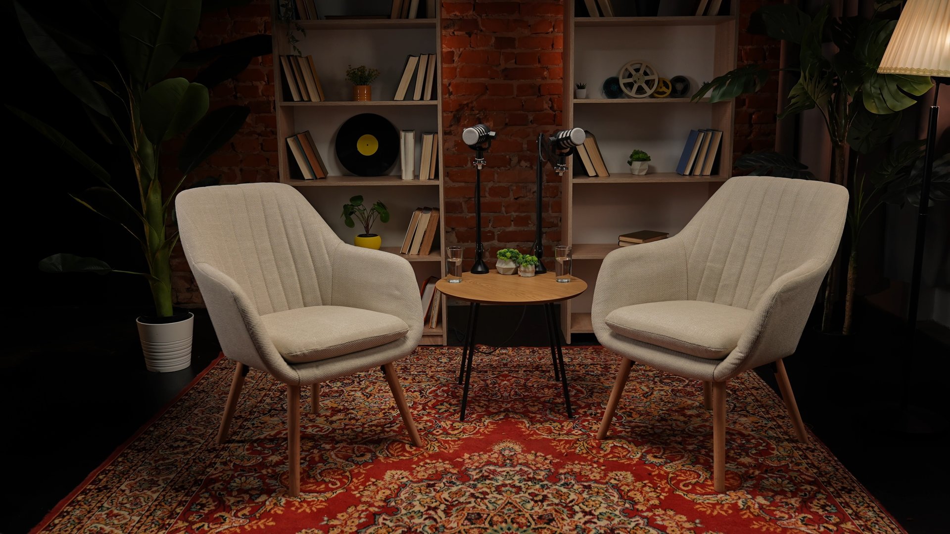 A stylish podcast studio setup featuring two modern armchairs, microphones, and a wooden table against a brick wall with bookshelves. Warm lighting creates an inviting atmosphere.