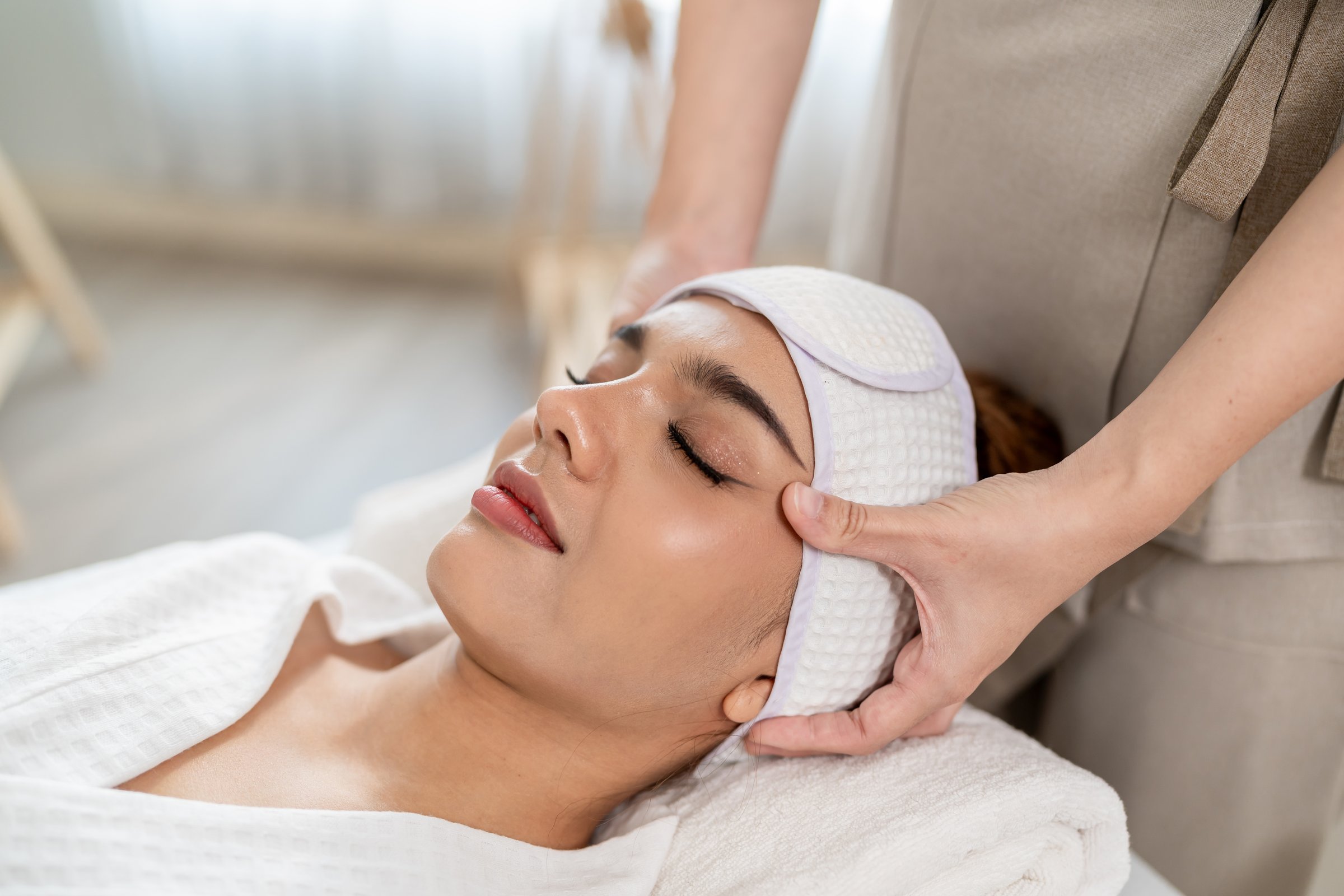Beautiful woman getting head and facial massage treatment at beauty spa. Attractive girl lying down on massage bed, getting relaxing physiotherapy service from masseuse for body care in spa clinic.