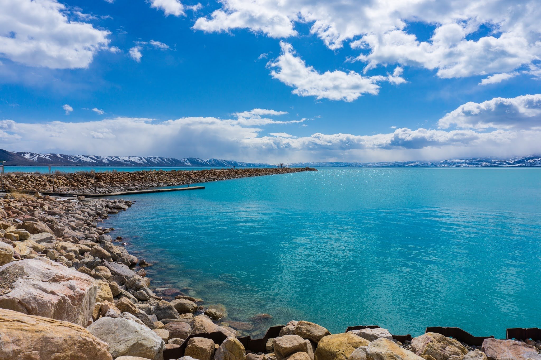 Rocky shoreline with turquoise water and distant snowy mountains under a blue sky with clouds.