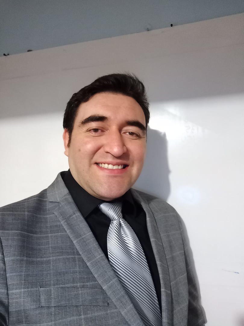 A man in a gray plaid suit and silver tie smiles while standing indoors against a plain white background.