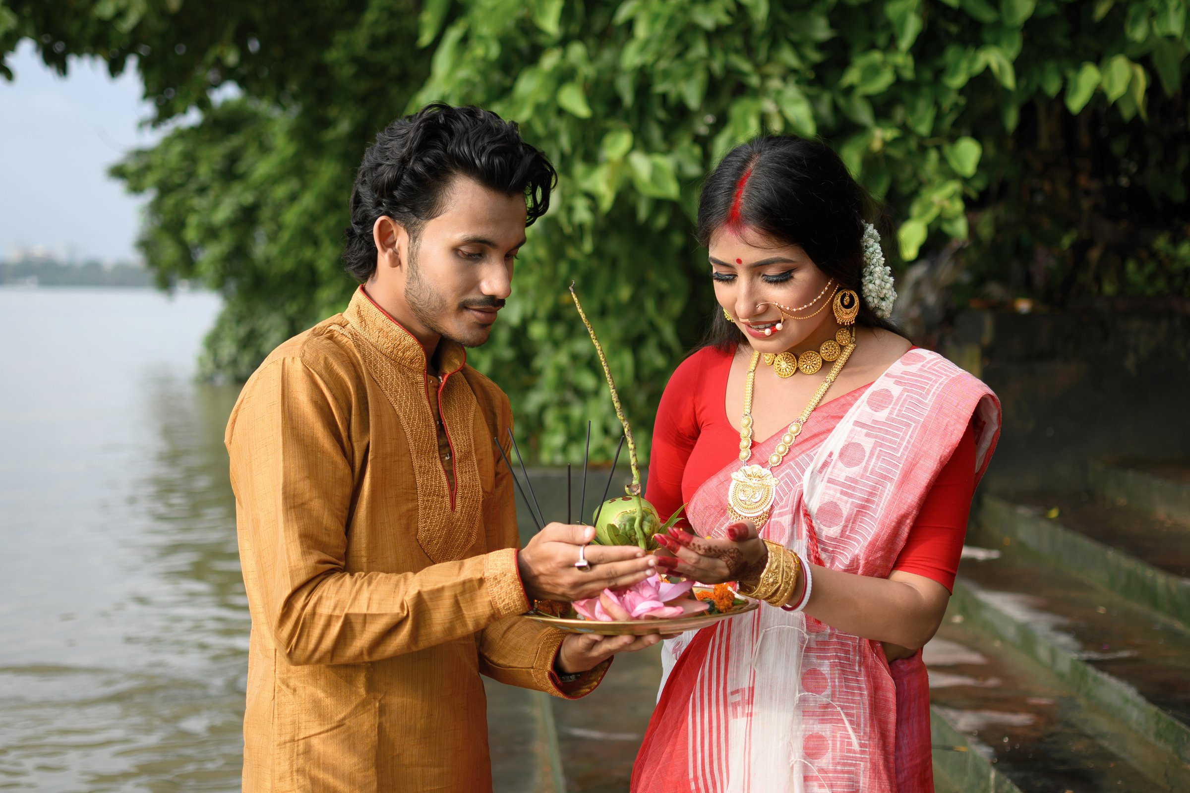 Portrait of a man in kurta pyjama and a woman in a traditional saree with gold jewellery, holding a plate of religious offerings in front of the river Ganges