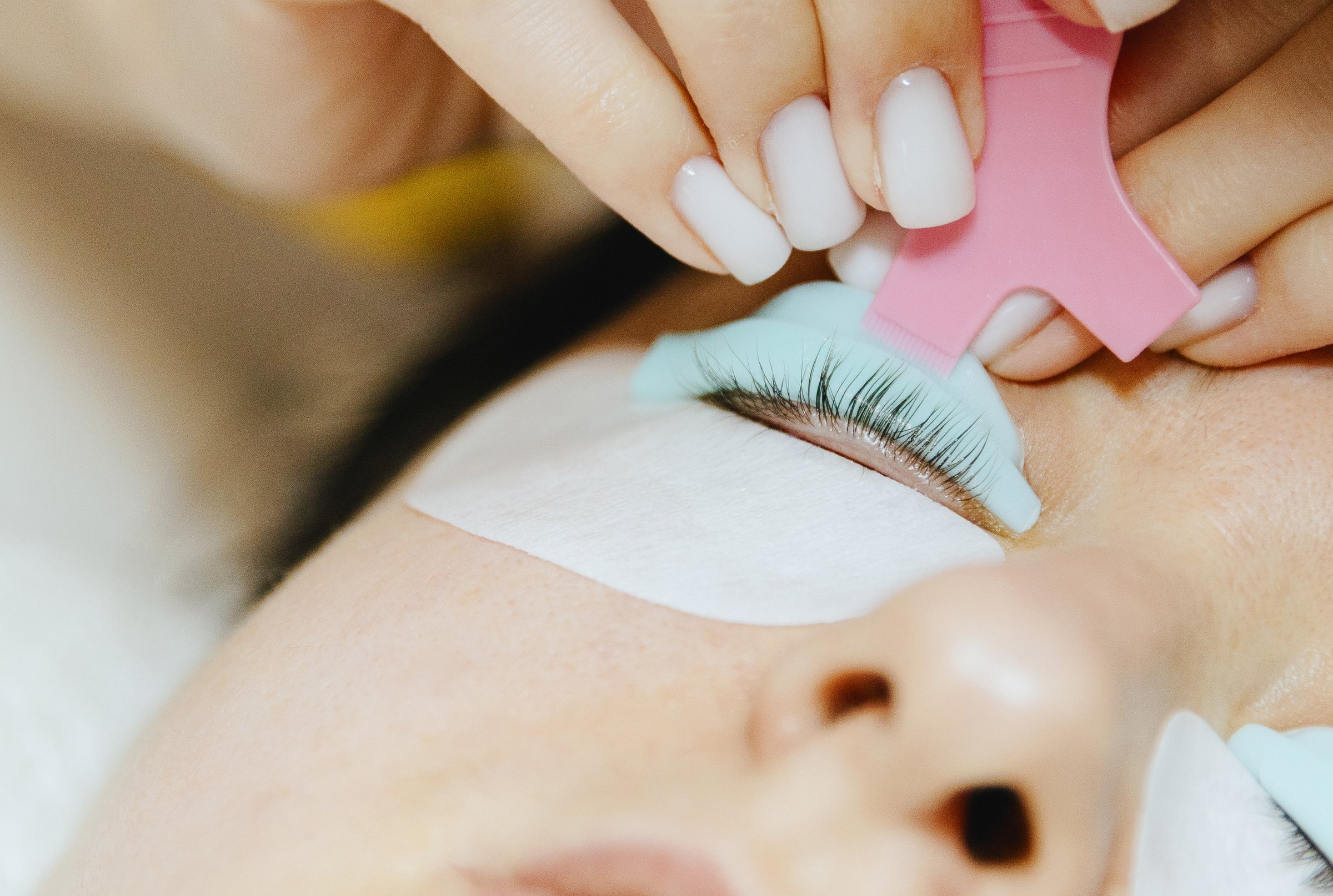 Portrait of a young unrecognizable Caucasian girl cosmetologist in a white coat who glues eyelashes onto a silicone mold using a pink plastic brush to a brunette woman lying on a cosmetology table in a home beauty salon for eyelash lifting, side view close-up with selective focus.