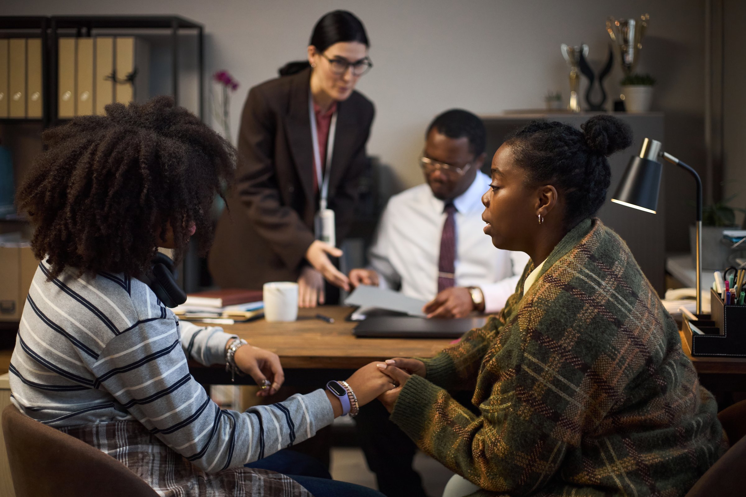 Middle aged Black woman holding hands with teenage Black girl, while sitting across desk from middle aged Caucasian woman and middle aged Black man discussing school matters in office
