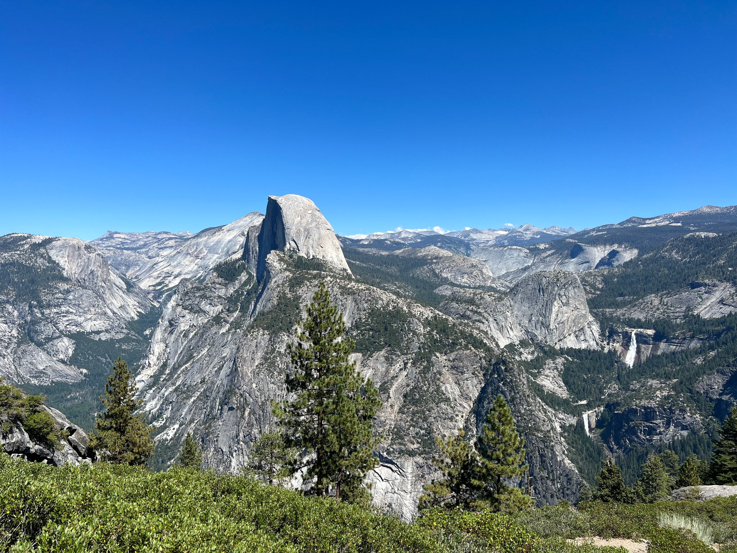 Panoramic view of Half Dome in Yosemite National Park with clear skies, forested foreground, and distant mountains.