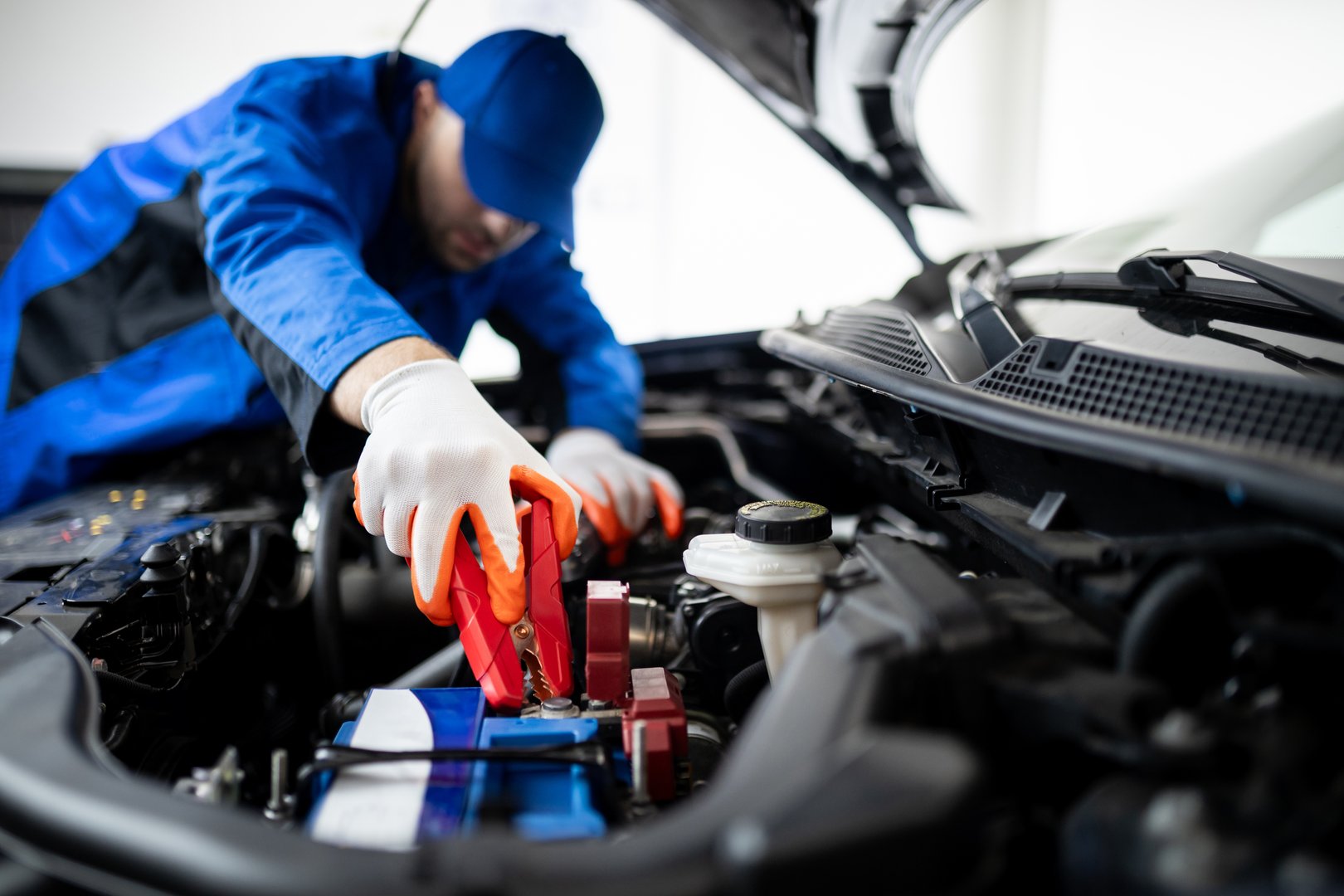 A technician in a blue uniform is focused on replacing a car battery in a well-lit workshop, using jumper cables to ensure proper connections.