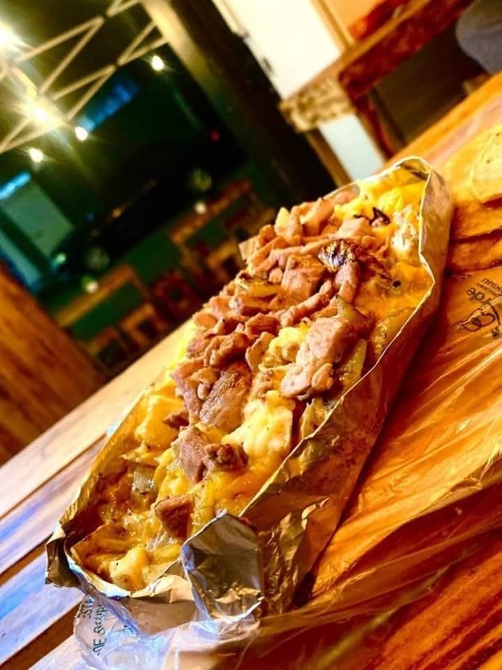 Loaded fries topped with grilled meat and cheese, served on foil, placed on a wooden table in a casual dining setting.