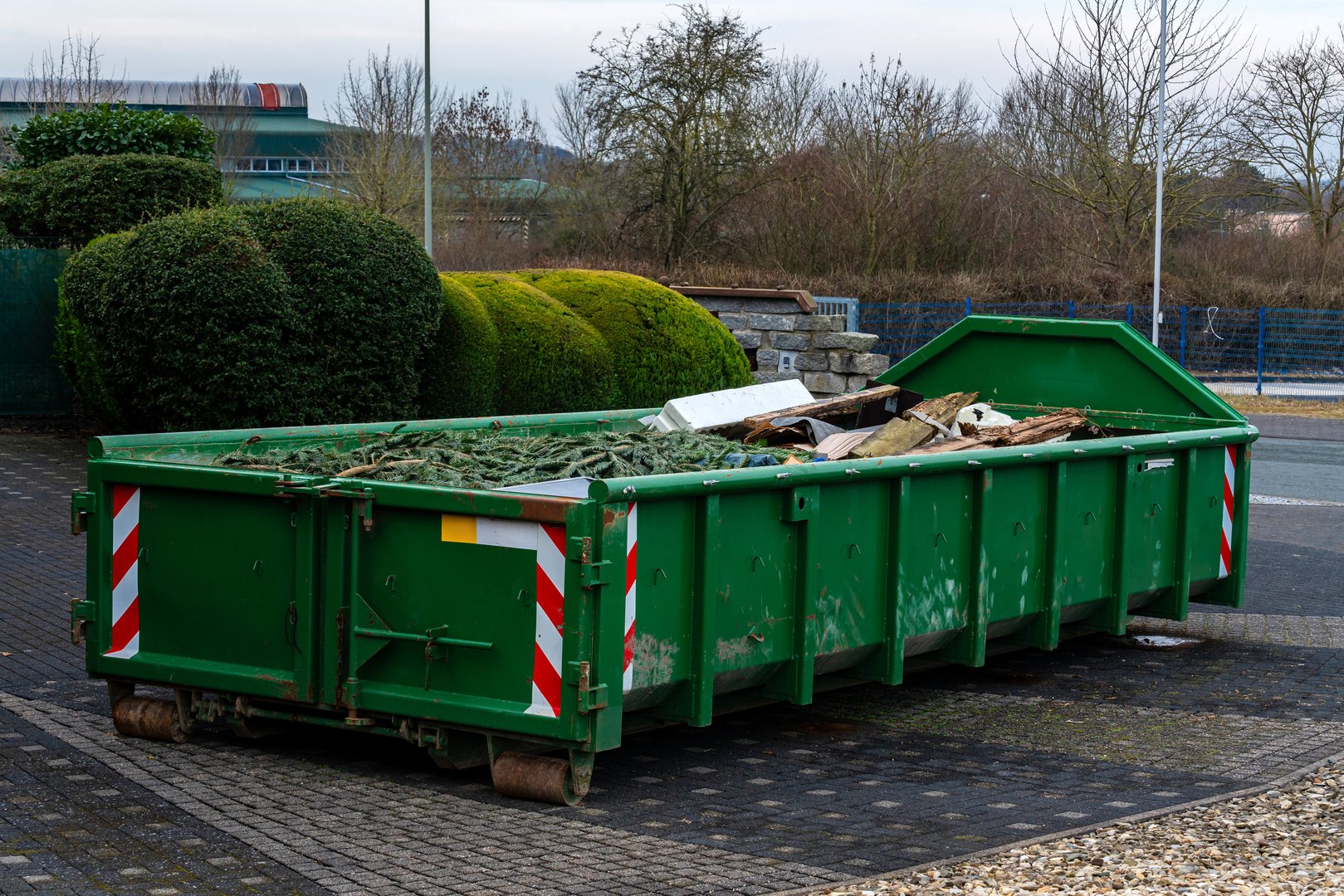A green dumpster is located in an urban setting, filled with various construction debris and materials.