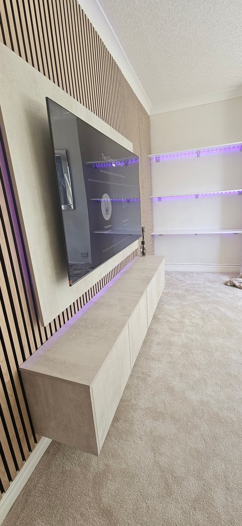Modern living room with a large wall-mounted TV, wooden paneling, floating cabinet, and shelves with purple LED lights.