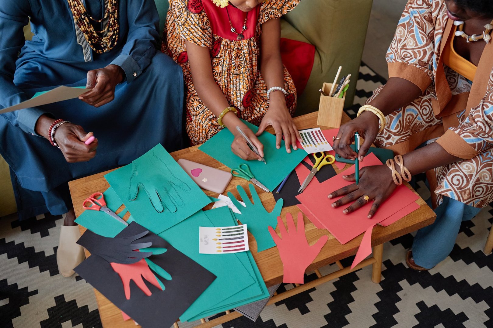 Black woman, Black man, and Black child creating colorful paper hand cutouts together at table, celebrating Kwanzaa with arts and crafts, hands actively drawing and cutting shapes