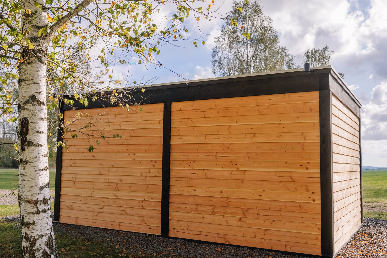 Modern wooden garden building with horizontal timber cladding and black trim, set in a natural landscape with a birch tree and cloudy sky in the background.