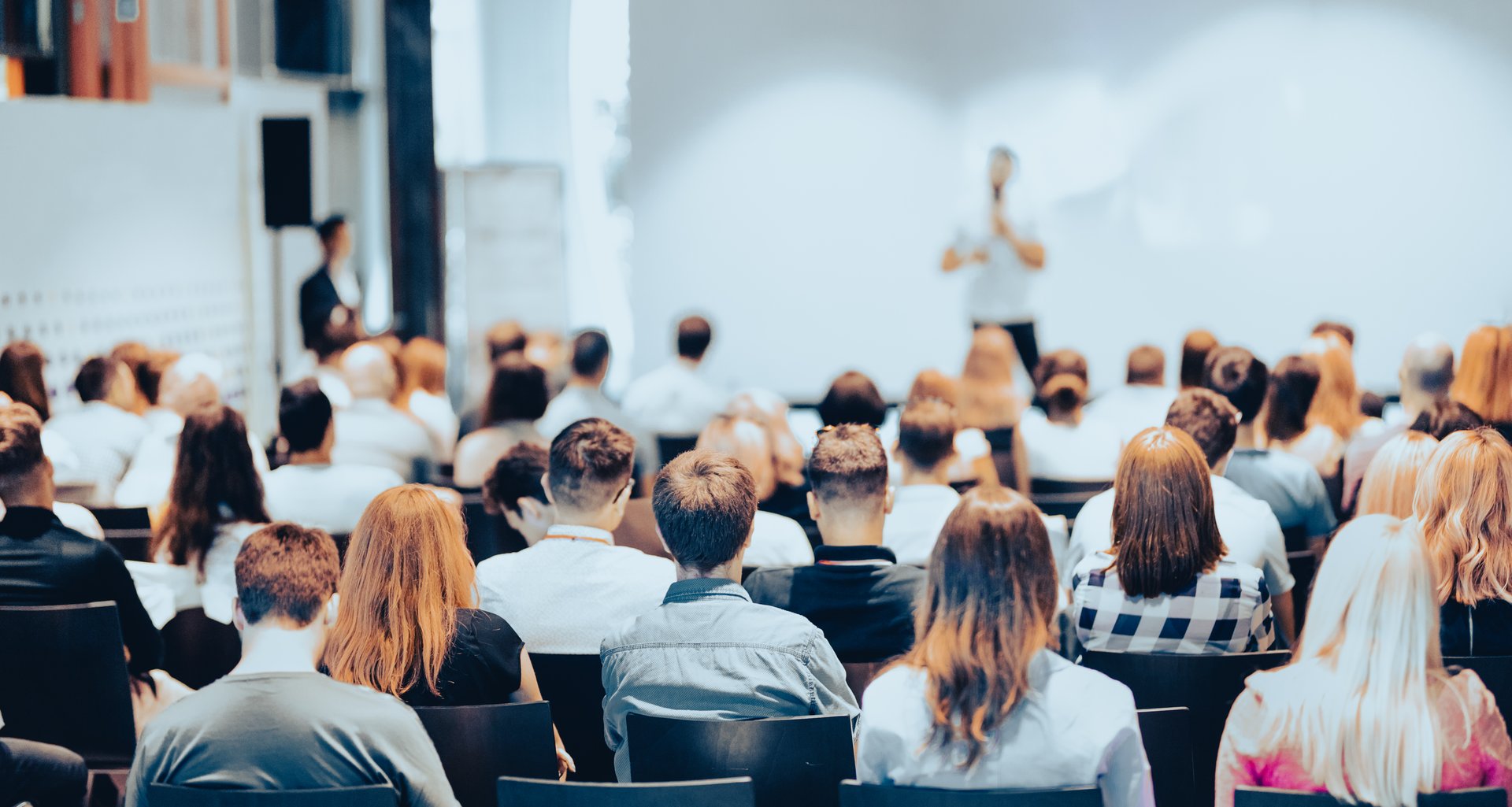 People seated in a conference room listening to a speaker addressing the audience from the front of the room.