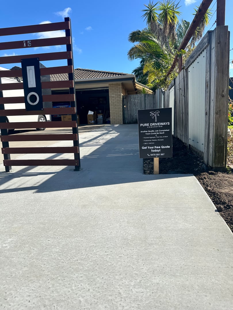 A clean concrete driveway leads to a garage. A sign for Pure Driveways is displayed near the entrance, offering service details.