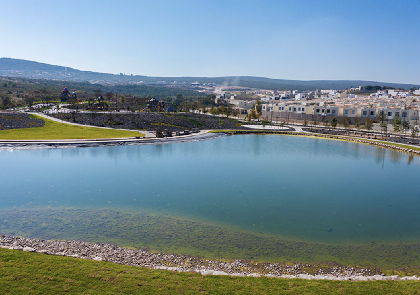 A calm lake with residential buildings in the background and hills under a clear blue sky.