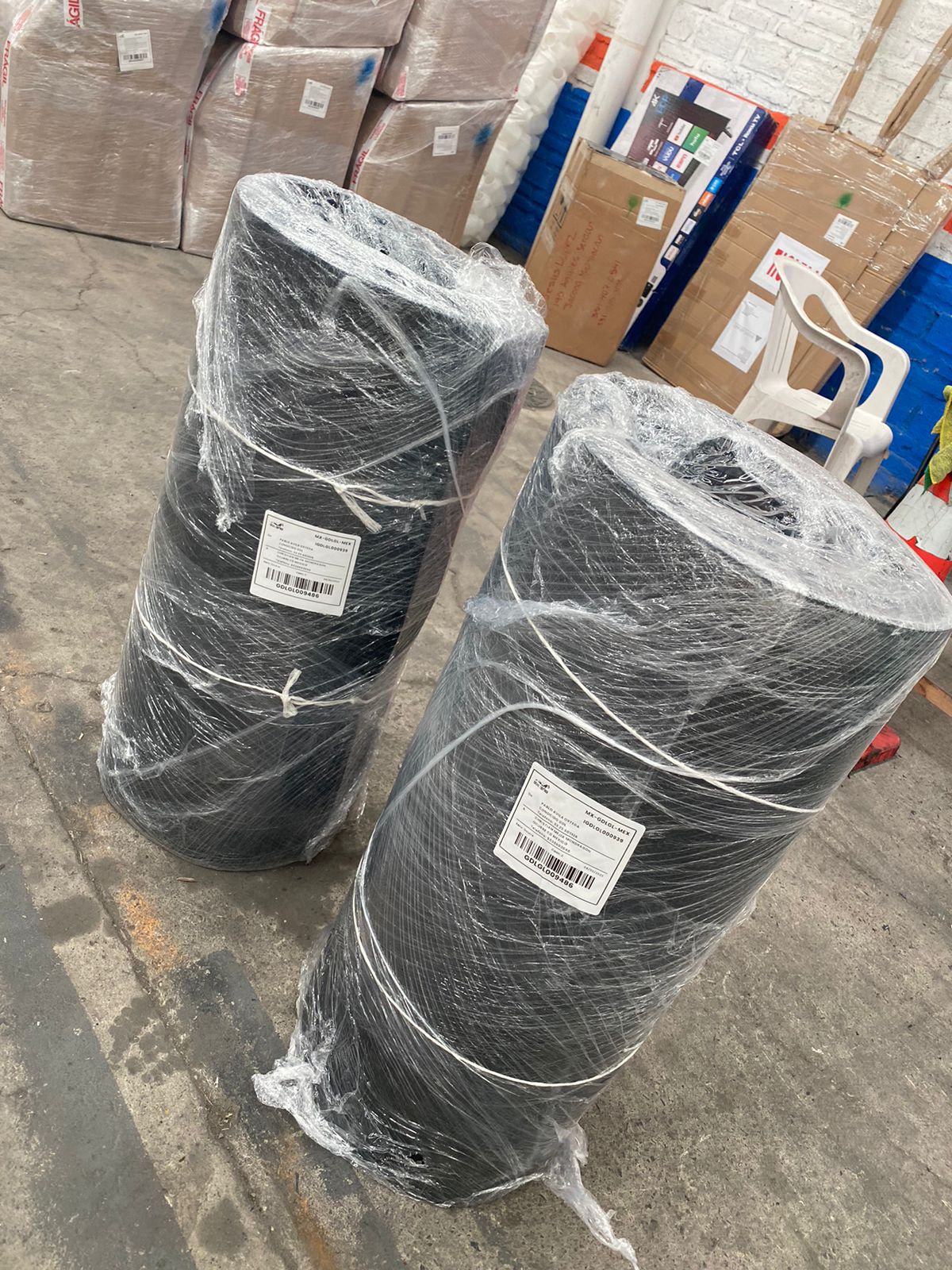 Two large black rolls wrapped in plastic, standing on a concrete floor with labels, surrounded by boxes and a plastic chair.
