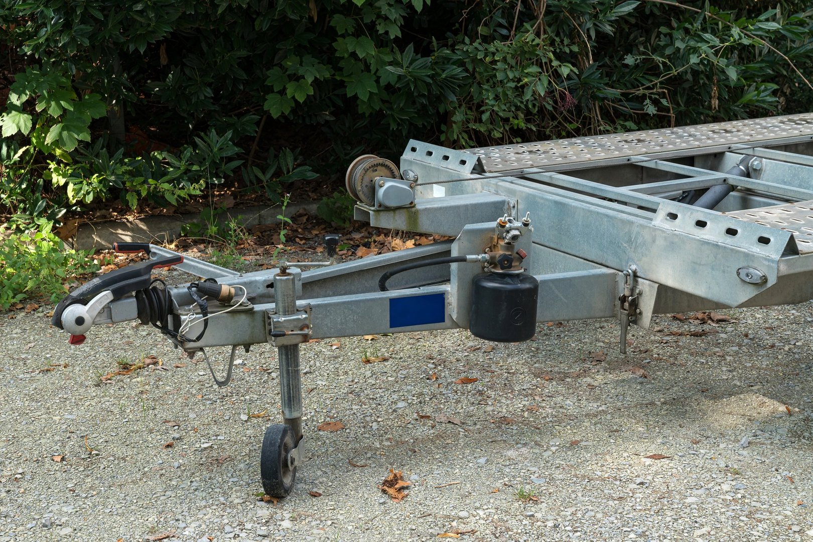 A close-up shot of a metal trailer hitch and chassis parked on a gravel path, with lush green foliage in the background.