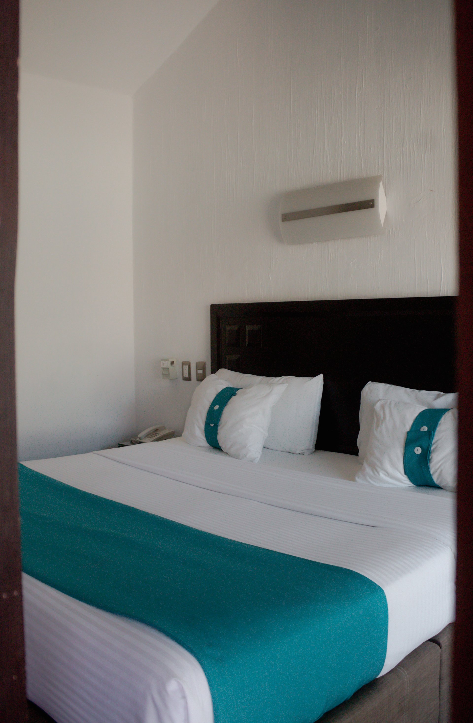 A neatly made hotel bed with white linens, teal accent runner, and matching pillows. Modern wall light above the bed.