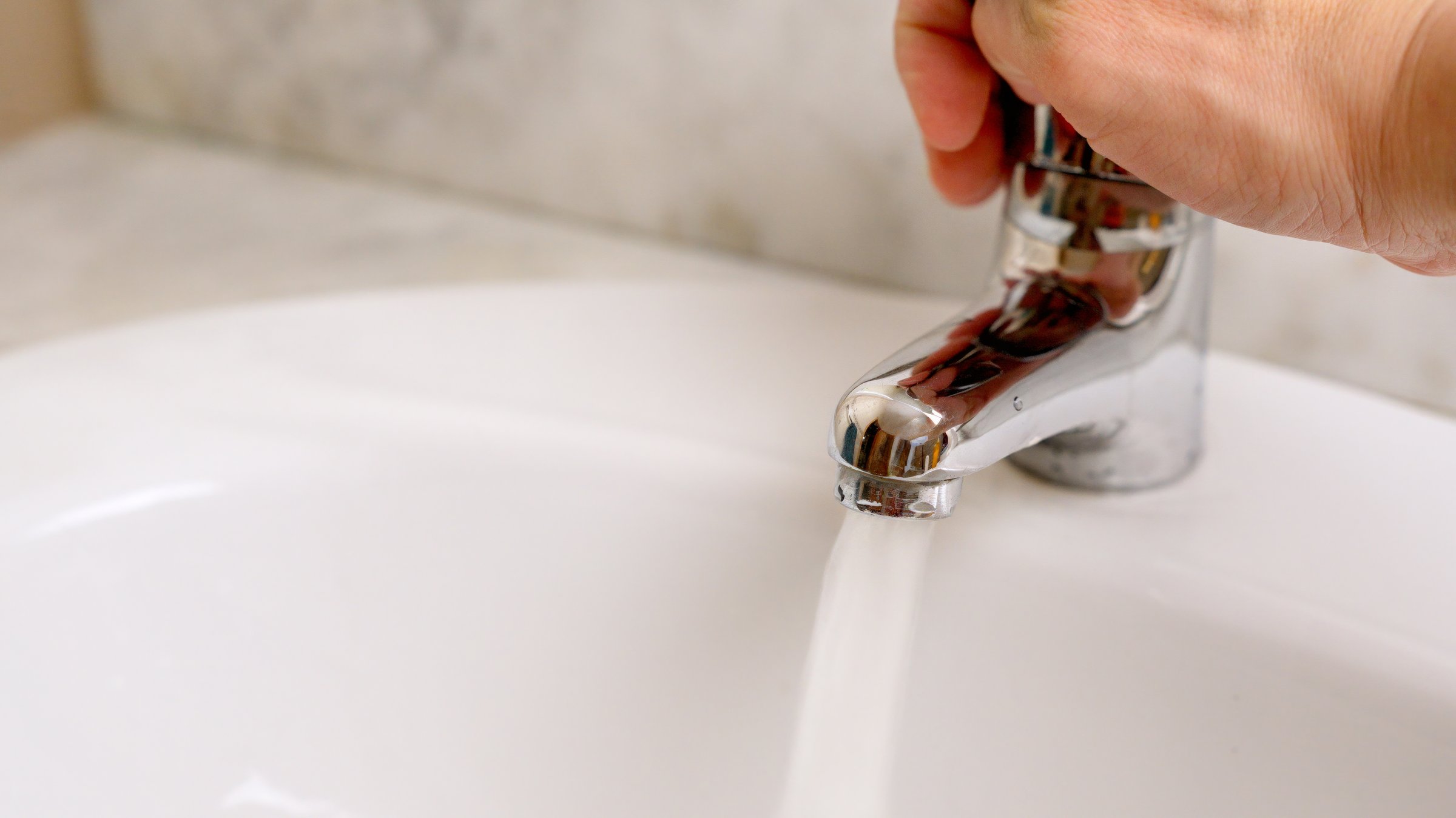 Hand turning on bathroom faucet with water pouring into sink, representing water conservation
