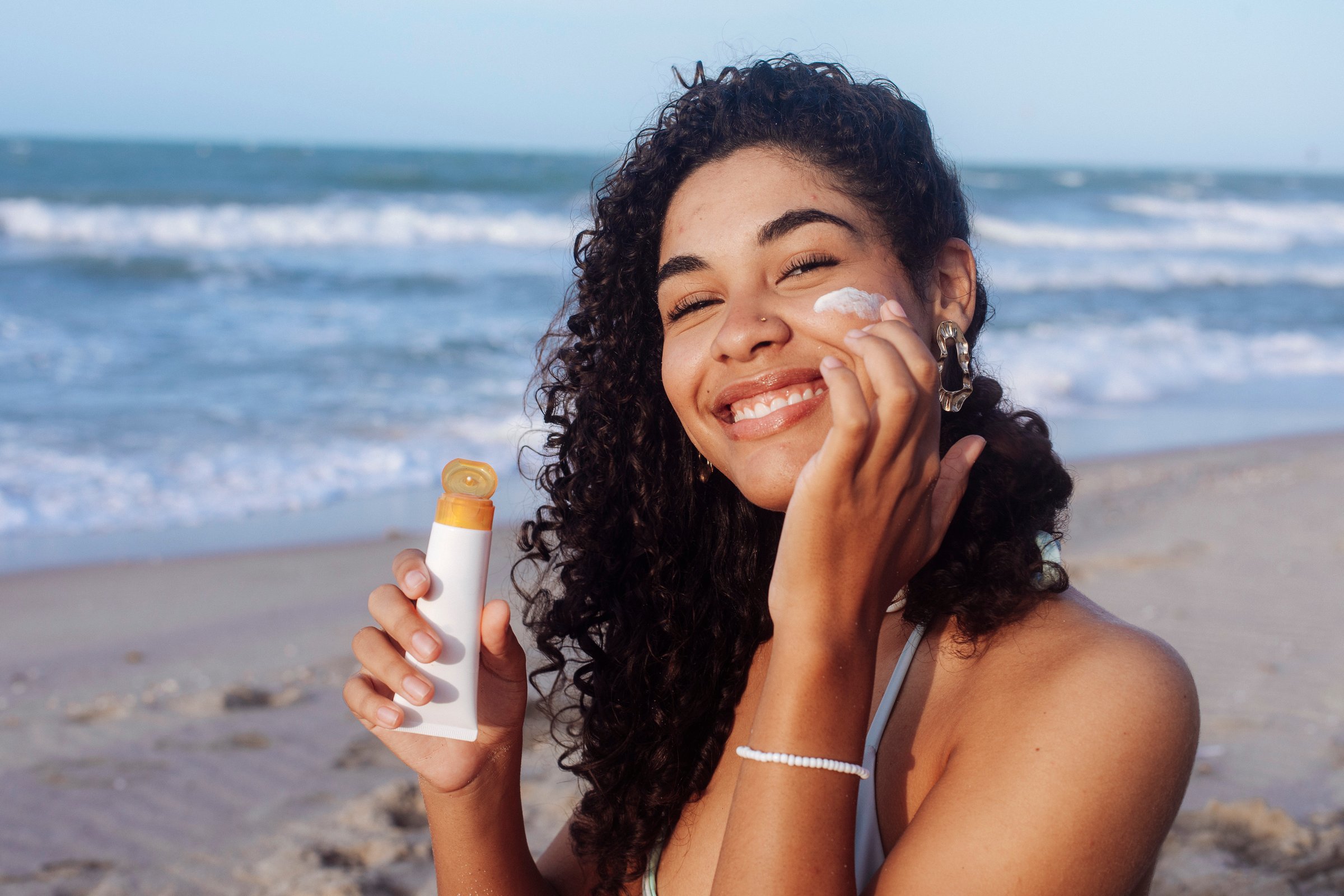 Close-up portrait of a young Latino-Hispanic woman with curly hair applying sunscreen to her face while enjoying a sunny day at the beach. She is smiling softly, wearing a light blue bikini, and protecting her skin under the warm sunshine, capturing a moment of self-care, natural beauty, and summer wellbeing.