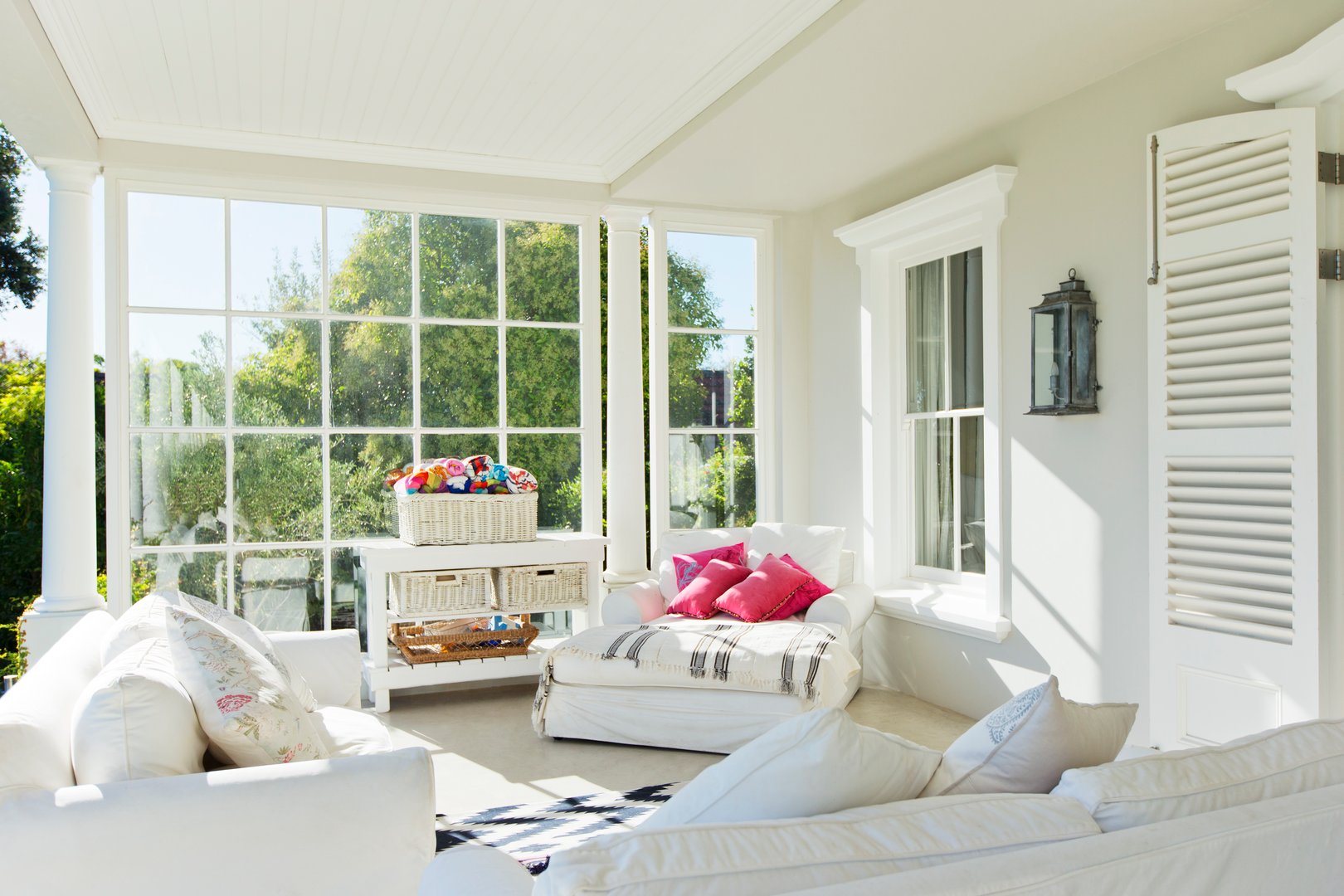 Bright sunroom with white sofas, colorful cushions, wicker baskets, large windows, and a garden view.