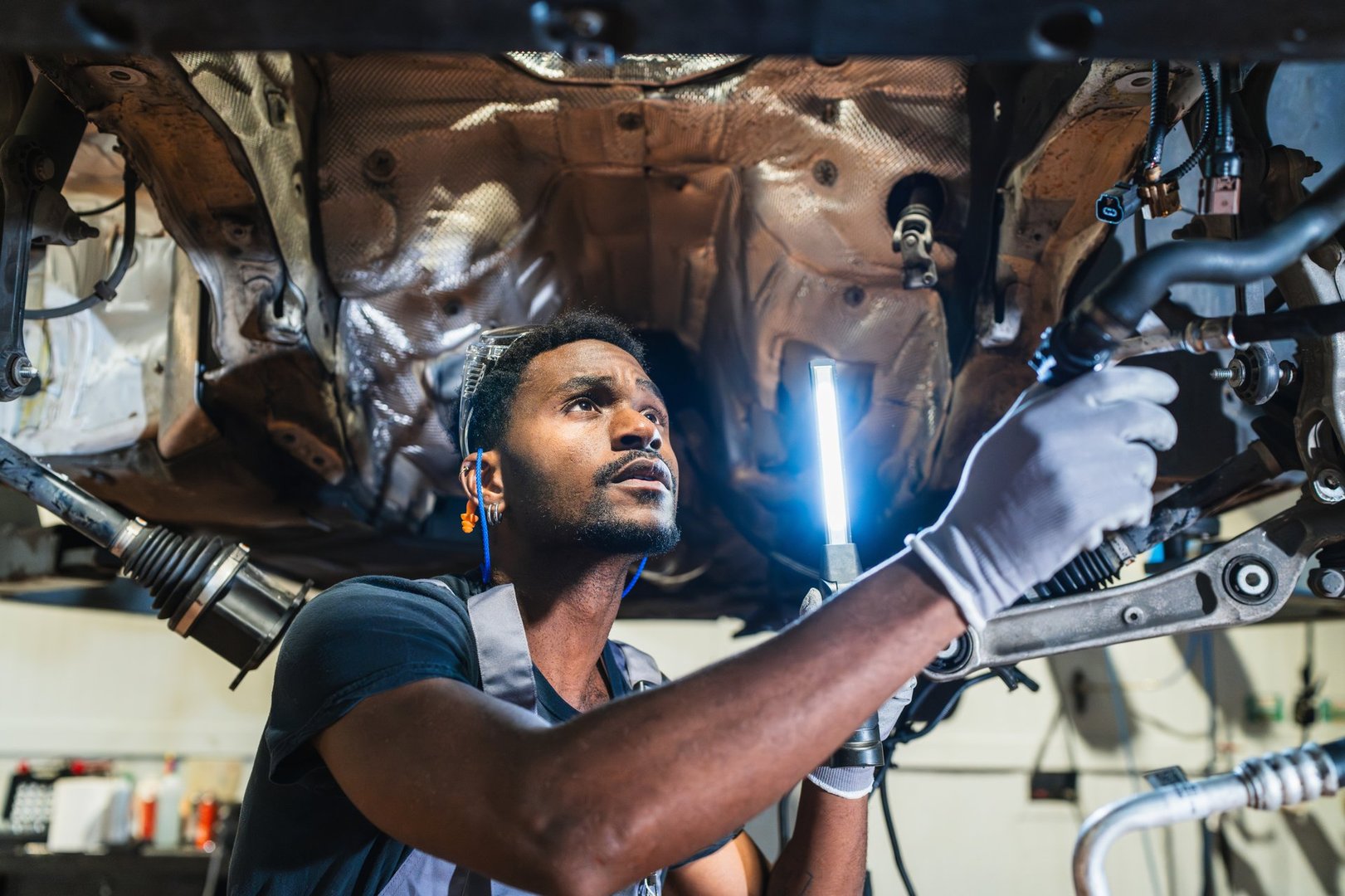 Young mechanic wearing gloves using flashlight inspecting underside of vehicle in auto repair shop