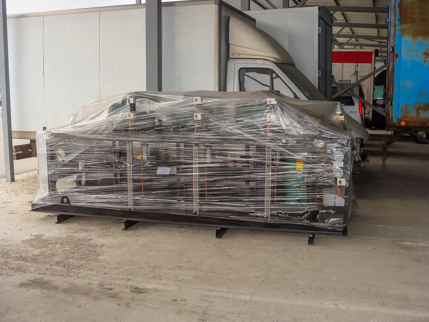 Industrial equipment, wrapped in protective plastic, sits on a metal skid inside a warehouse, ready to be shipped or stored.