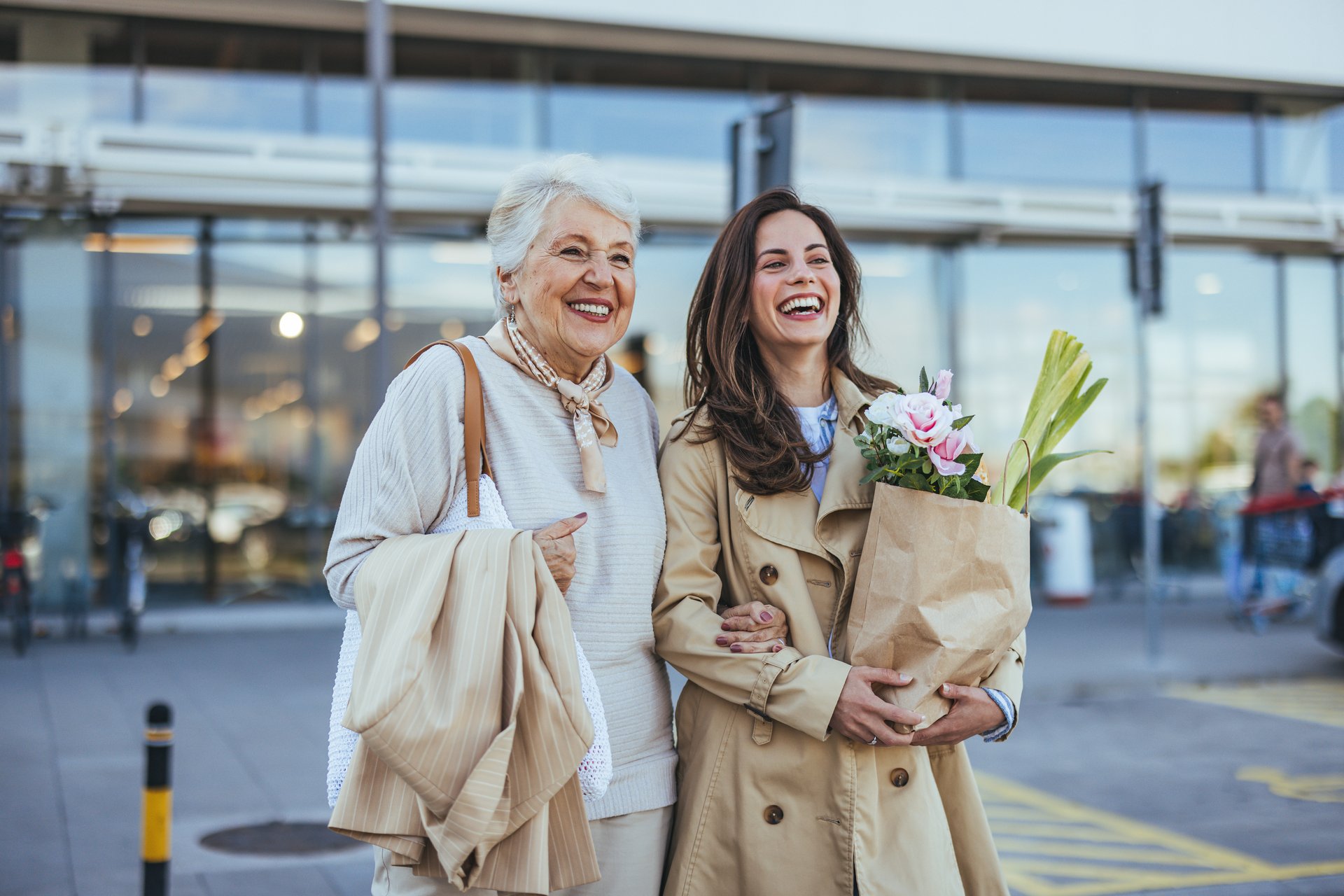 A cheerful elderly woman and her adult daughter enjoying a shopping trip together. They are smiling while holding a bouquet of fresh flowers outside a store.
