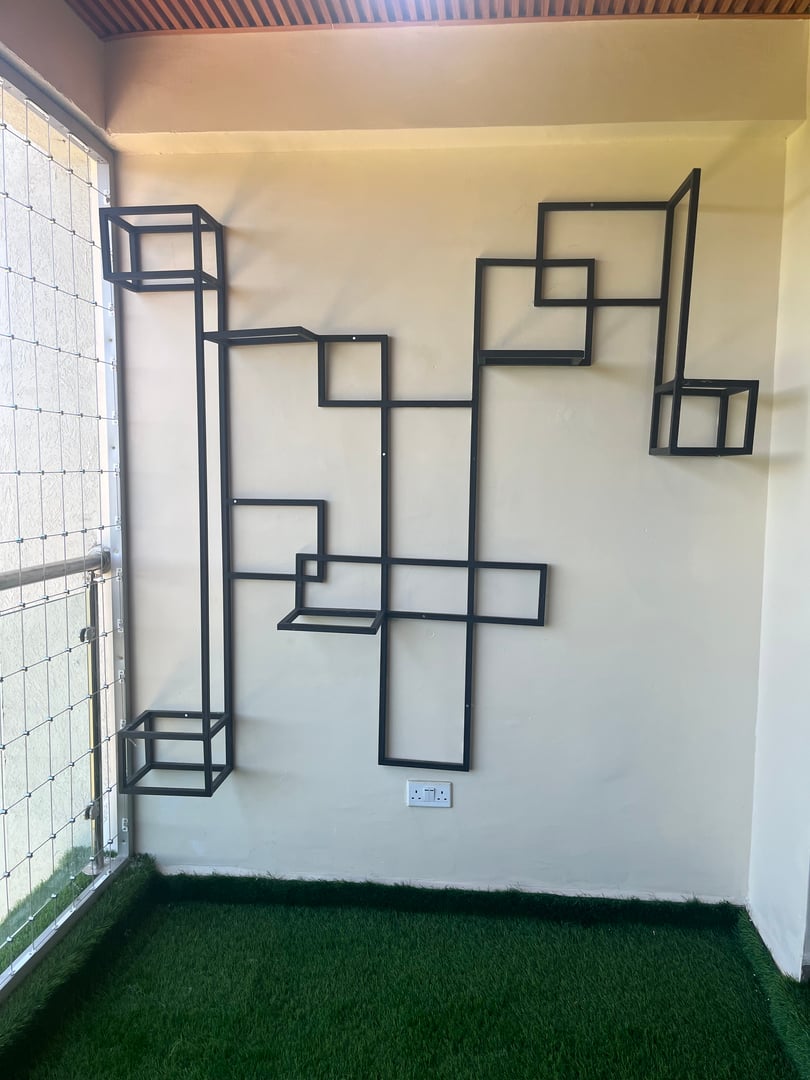 Geometric metal wall shelf with overlapping rectangular frames against a white wall, placed on artificial grass flooring.