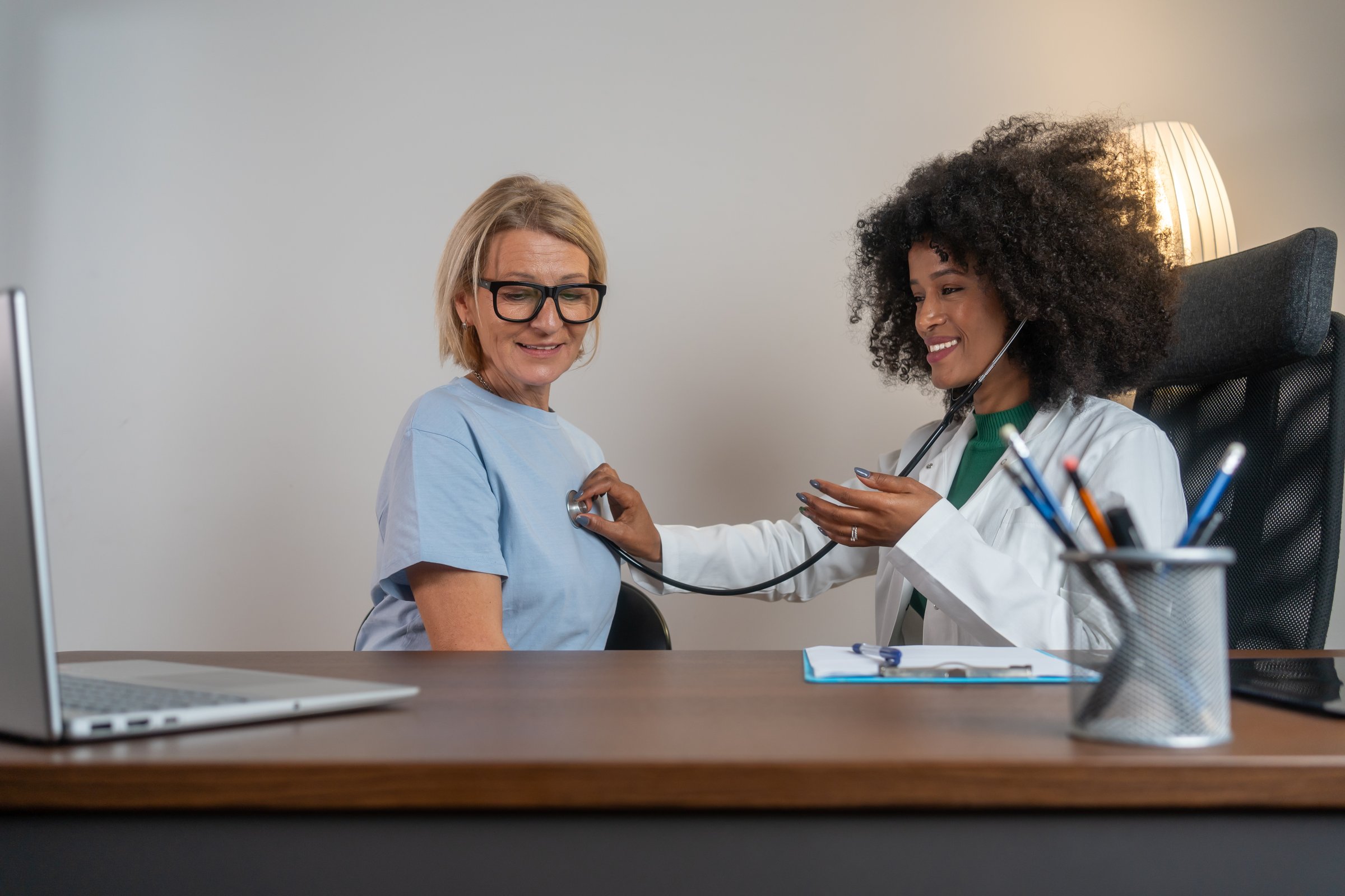 Young african female doctor listen to female patient heart chest with stethoscope at clinic meeting. Woman GP checkup examine woman client doctor listens to the patient's lungs and heartbeat.