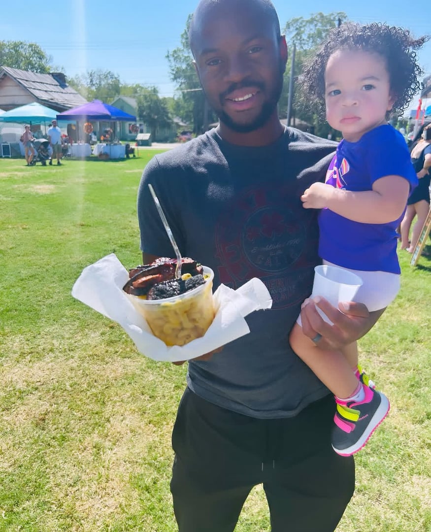 Man holding a bowl of food and a child at an outdoor event on a sunny day.