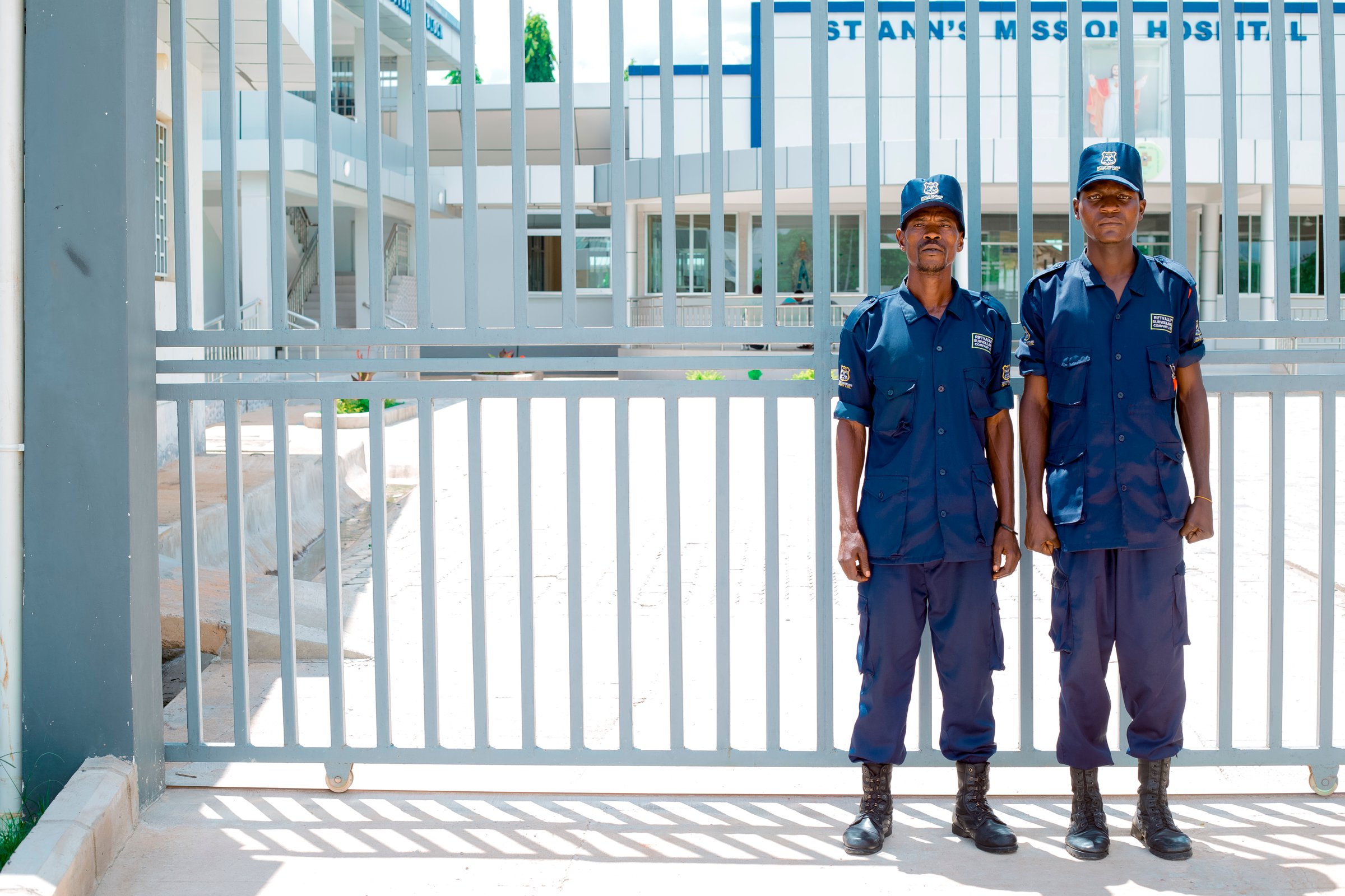 Two security guards in blue uniforms stand in front of a gated entrance to a modern building on a sunny day.