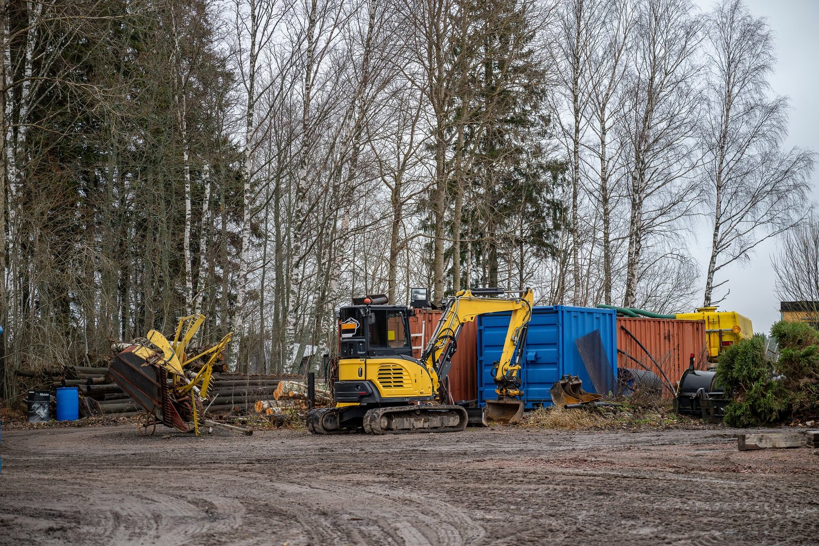 Yellow excavator parked near colorful containers and machinery on a dirt-covered industrial site