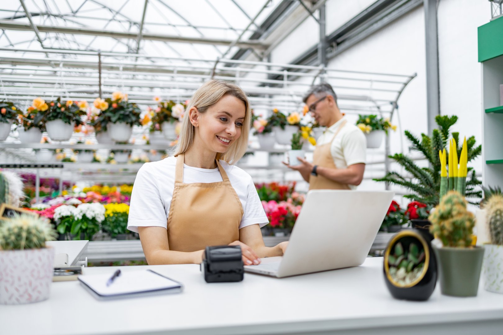 A smiling Caucasian florist uses a laptop behind a sale table, with another employee visible in the background. The shop is filled with flowers and plants.