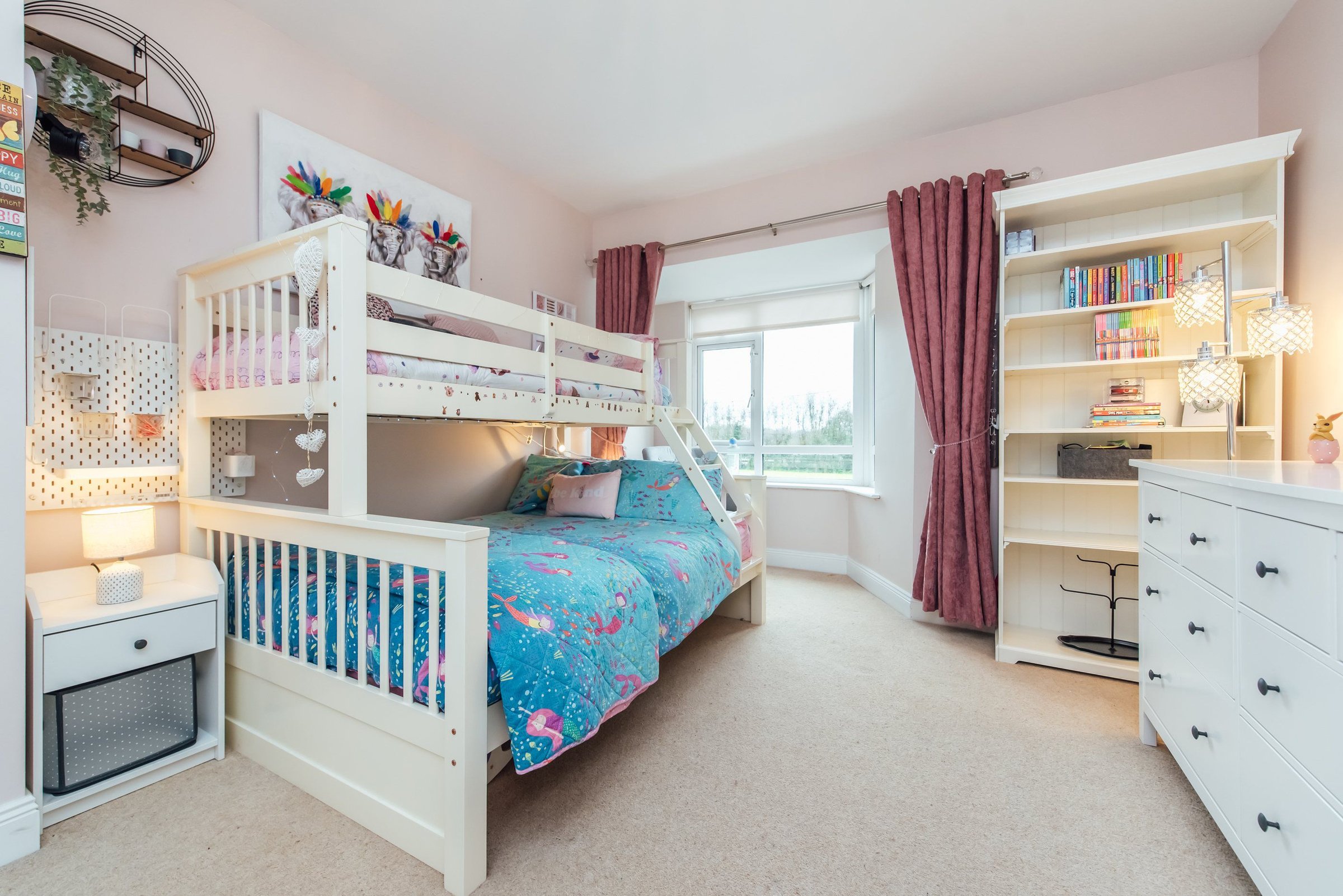 A bright childrens room with twin beds, colorful bedding, a shelf of books, and decorative lights.