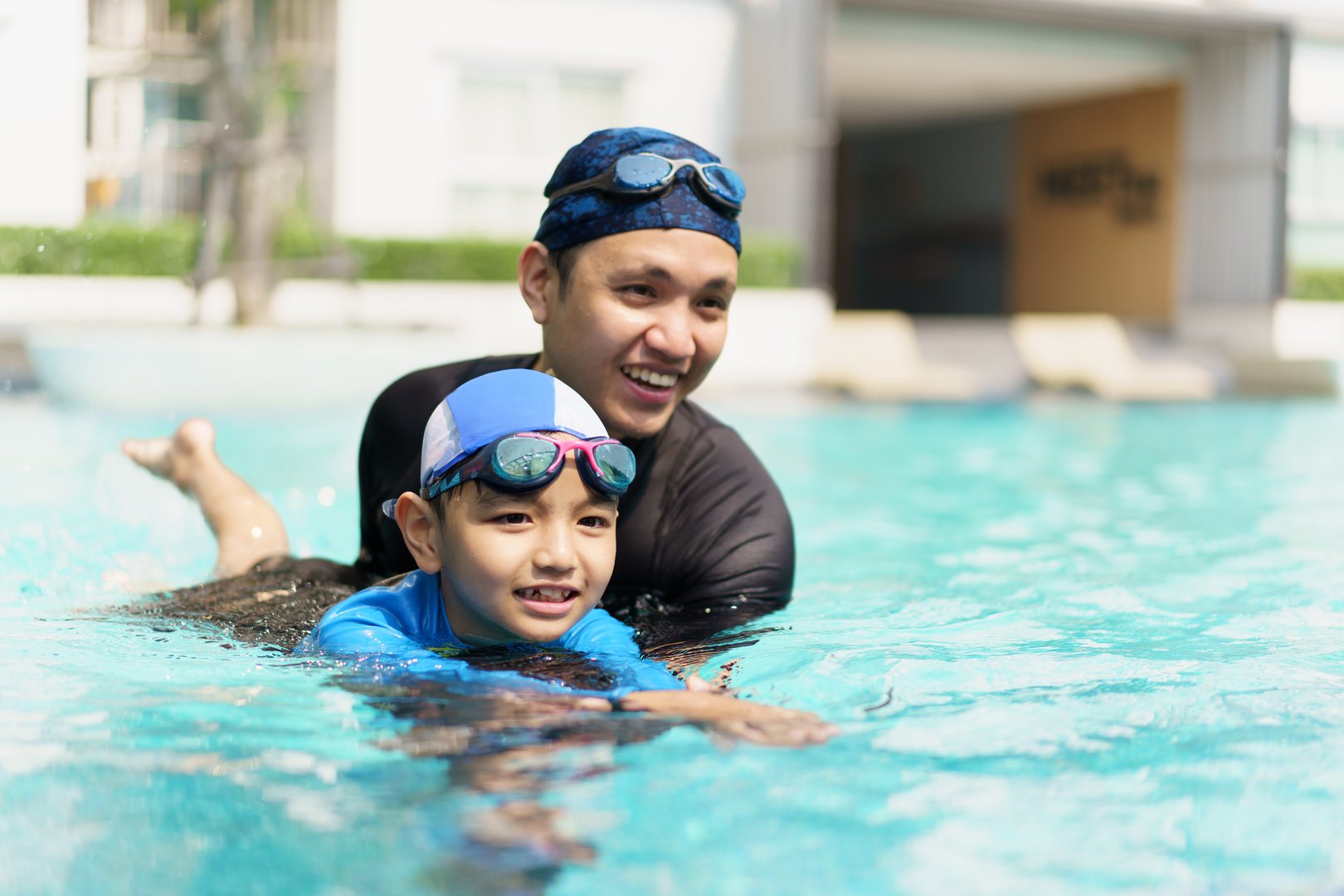 Happy cheerful Asian father and son playing and swimming together in the swimming pool, a boy enjoy playing a water with his dad. Summer and vacation activities in family.
