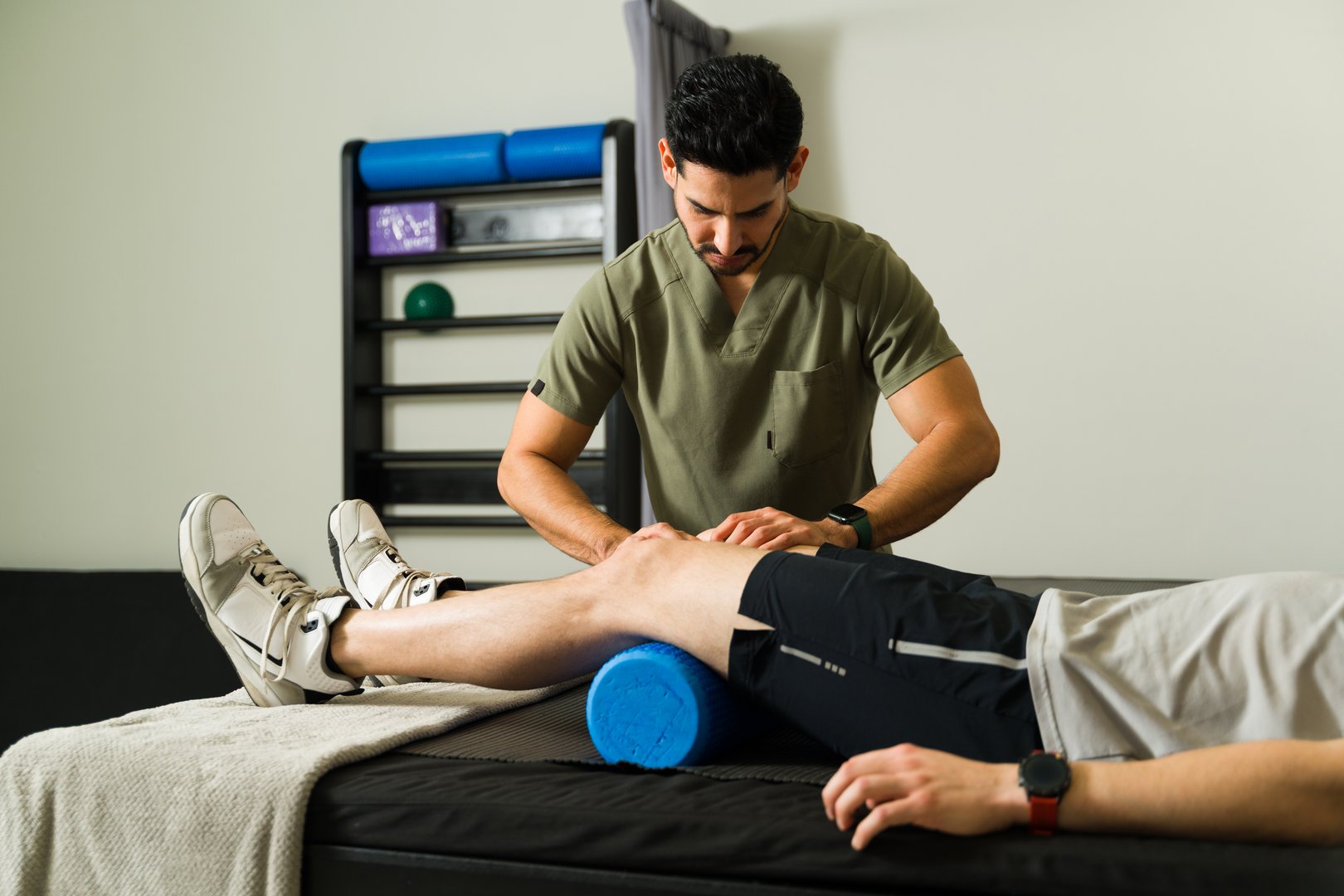 Latin american physiotherapist massaging athlete's knee with foam roller during physical therapy session in modern rehabilitation center