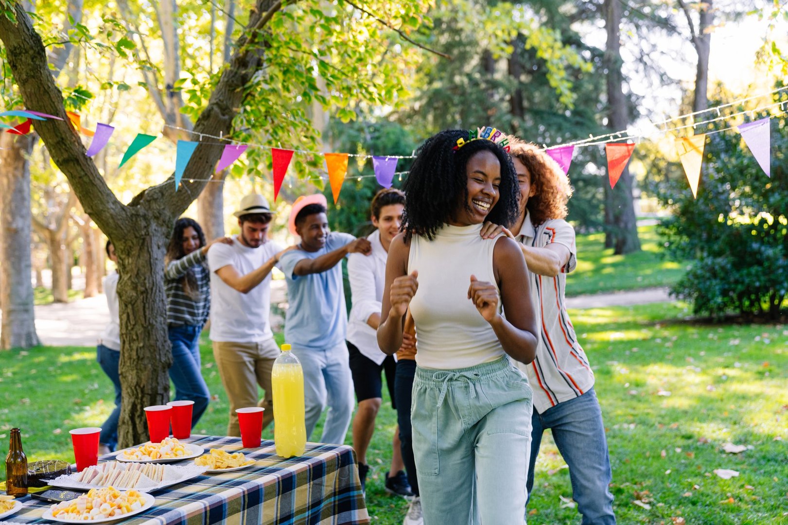 Young adults dancing joyfully in a conga line, celebrating a vibrant birthday party in the park, surrounded by friends and sunshine
