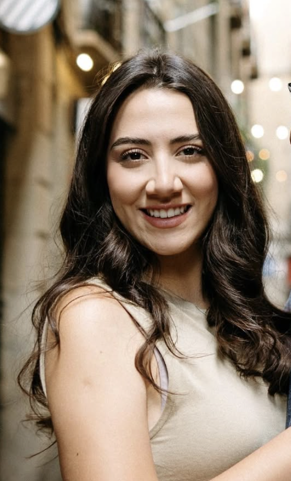Smiling woman with long dark hair in a sleeveless beige top, standing in a street with blurred lights in the background.