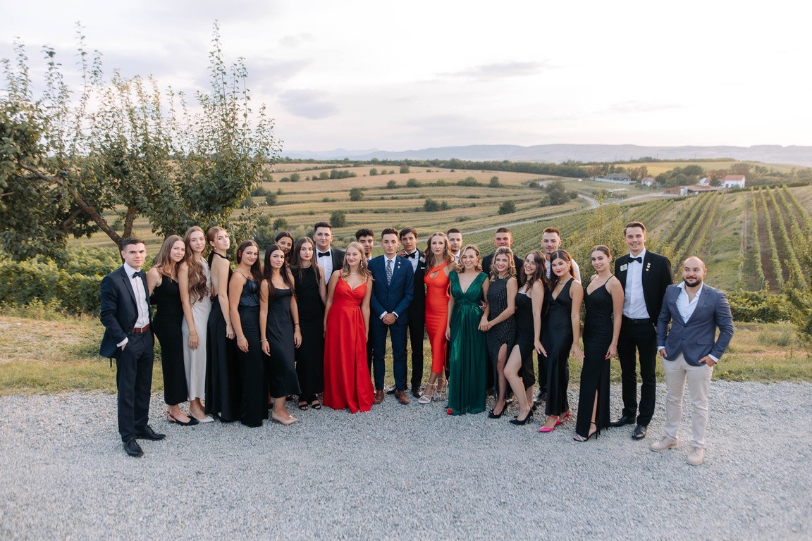 A group of elegantly dressed people posing outdoors on a gravel path, with vineyards and hills in the background.