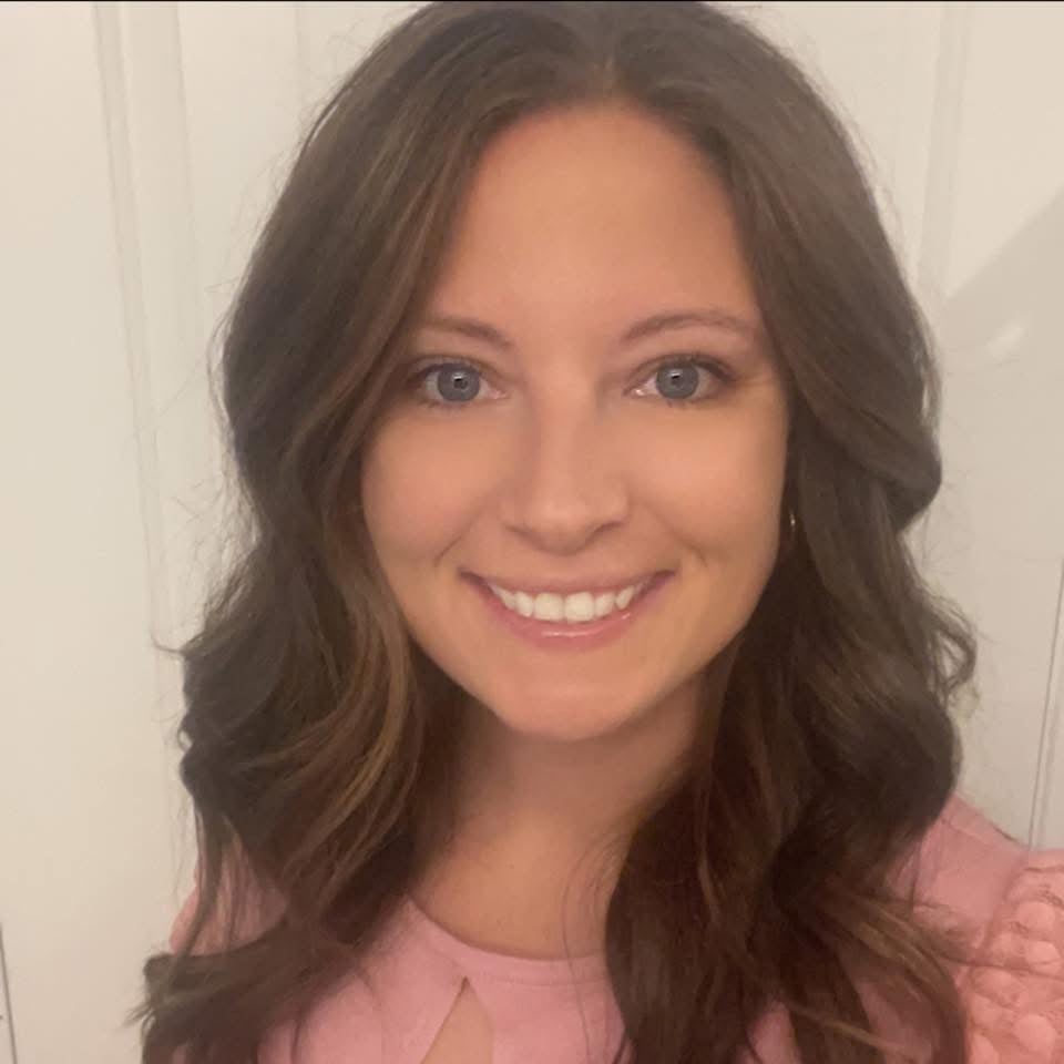 Smiling woman with long brown hair in a pink top, standing in front of a light-colored background.