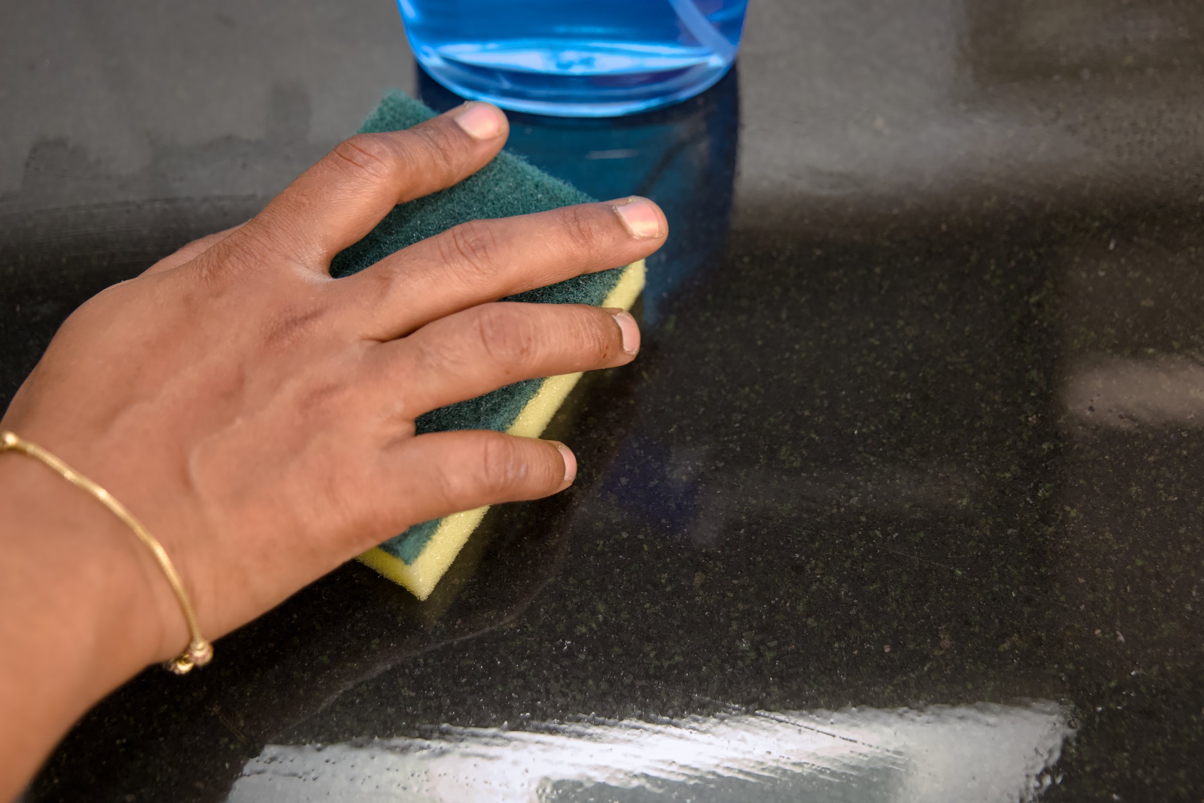 A hand diligently wipes a black granite countertop with a cleaning sponge.