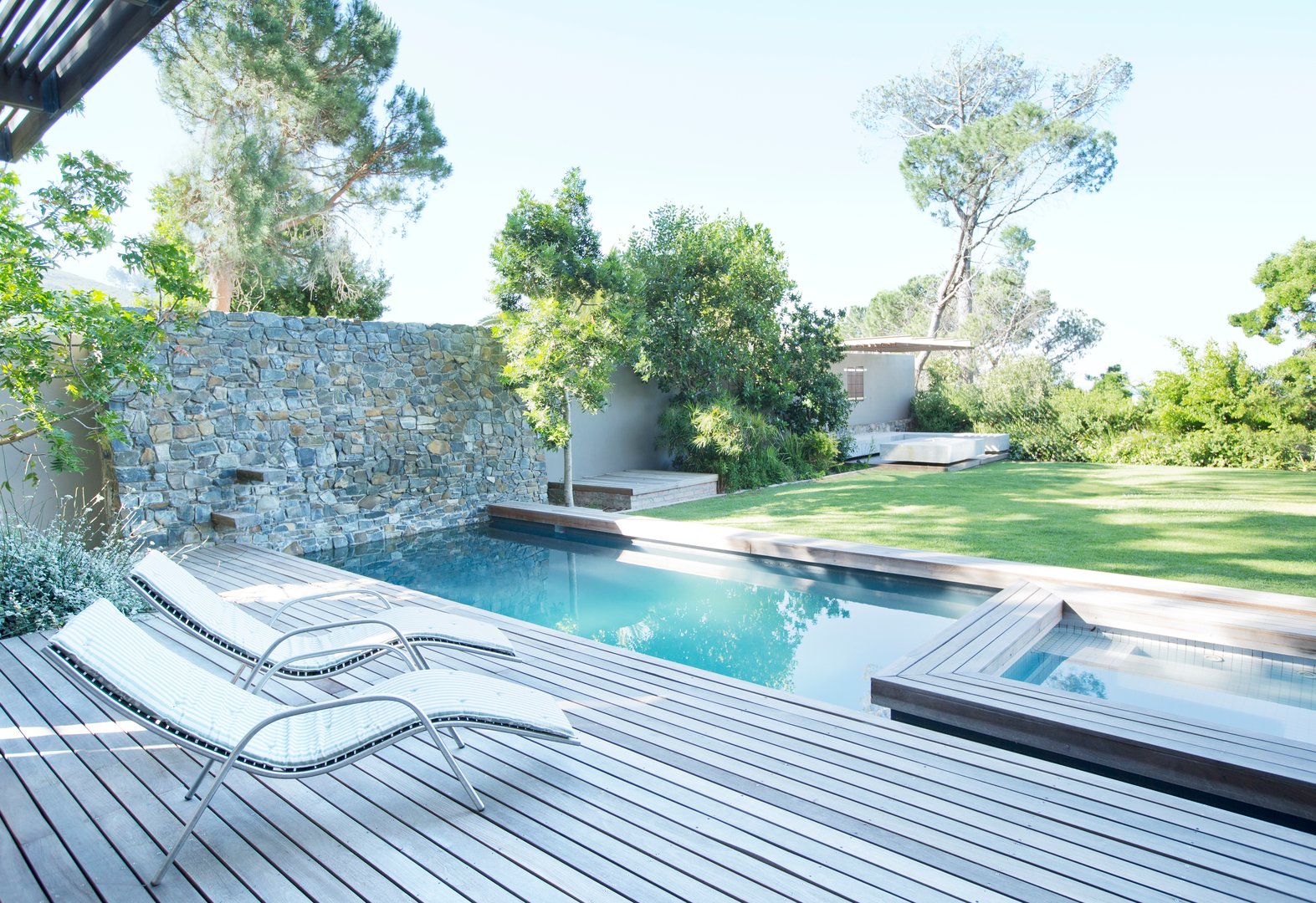 Modern patio with wooden deck, lounge chairs, rectangular pool, and lush garden backed by a stone wall and tall trees.
