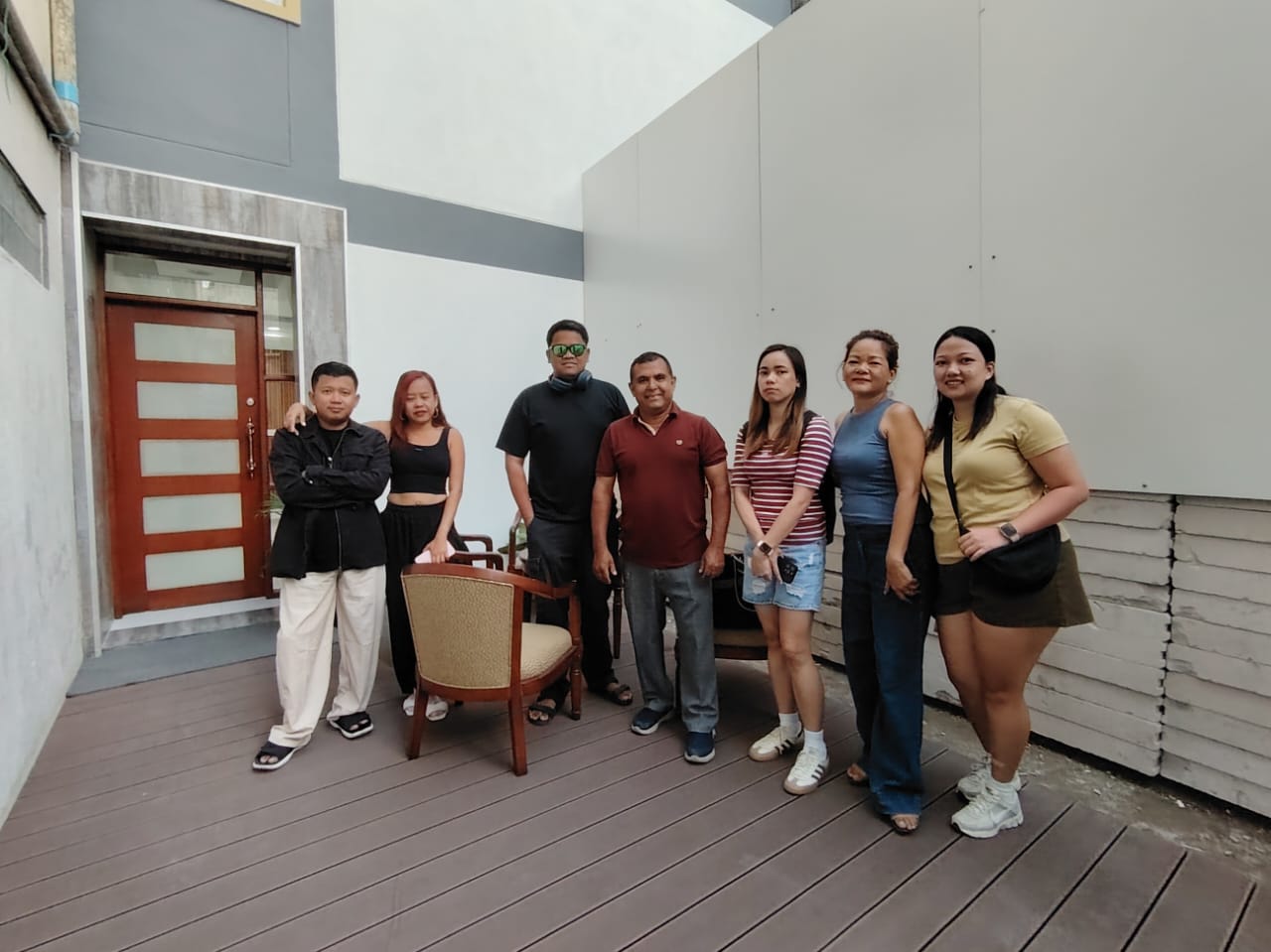 Group of seven people posing casually on a patio, with a chair and a modern building in the background.